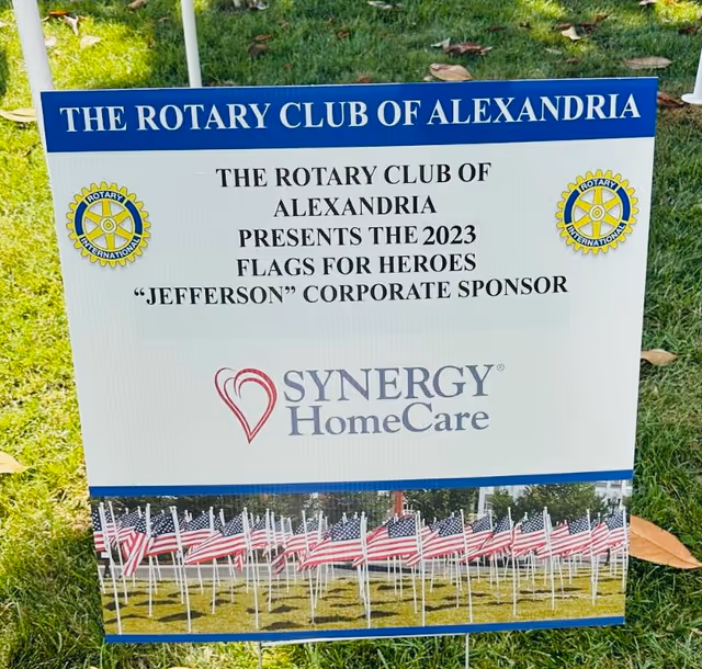 Outdoor sign shows Synergy HomeCare logo with Rotary Club banners and American flags.