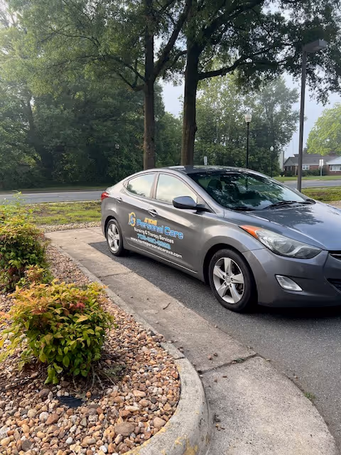 Gray branded car labeled At Home Personal Care Services parked beside landscaped curb.