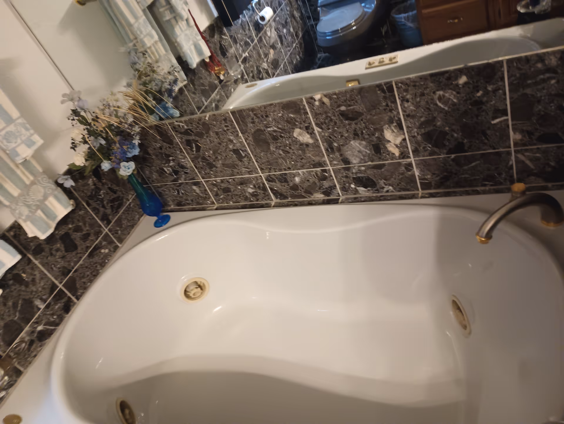White bathtub with dark marble tiles, flowers, and a mirror in a bathroom.