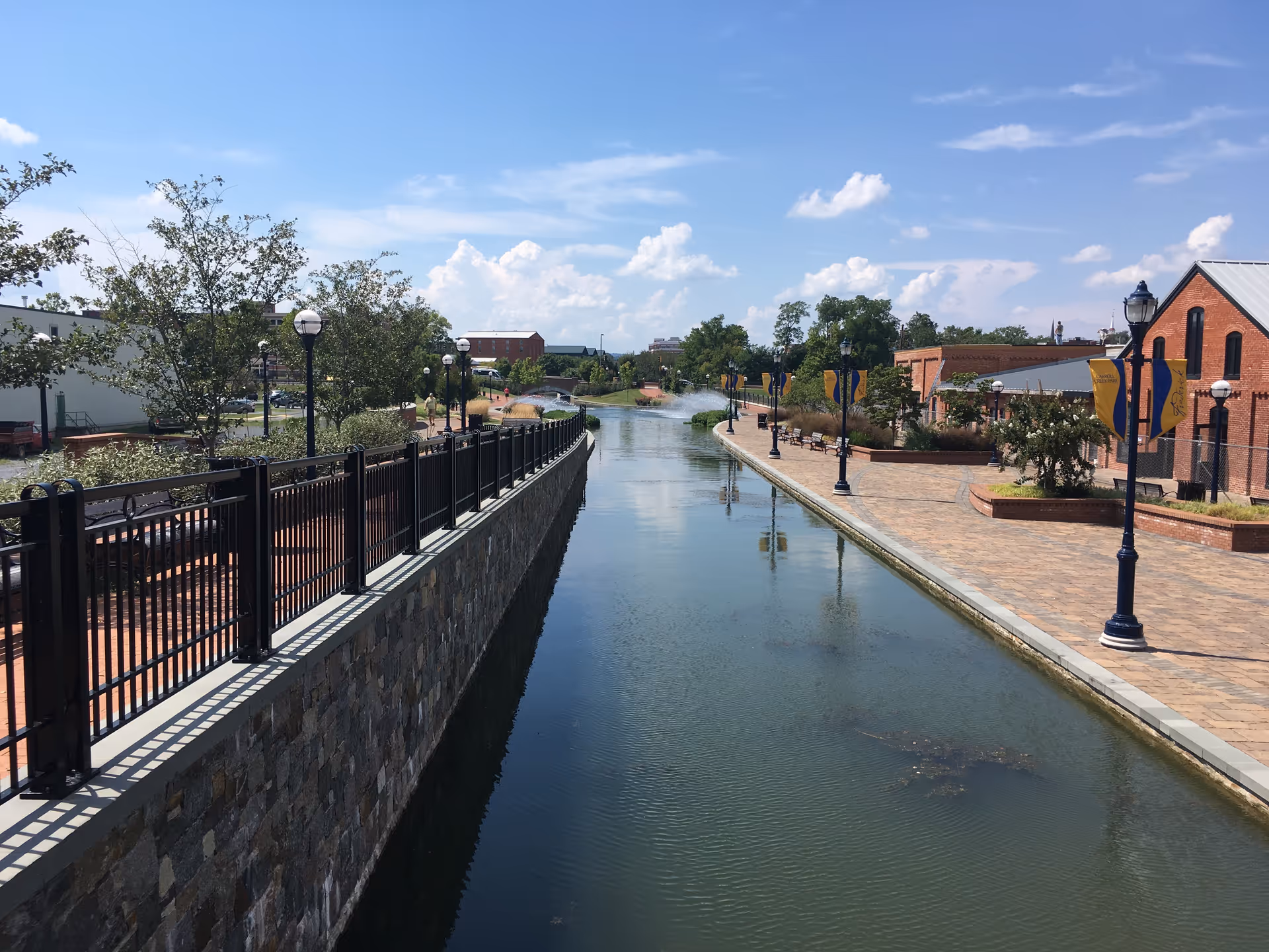 Outdoor canal with railing, brick walkway, lamps, and trees on a sunny day.