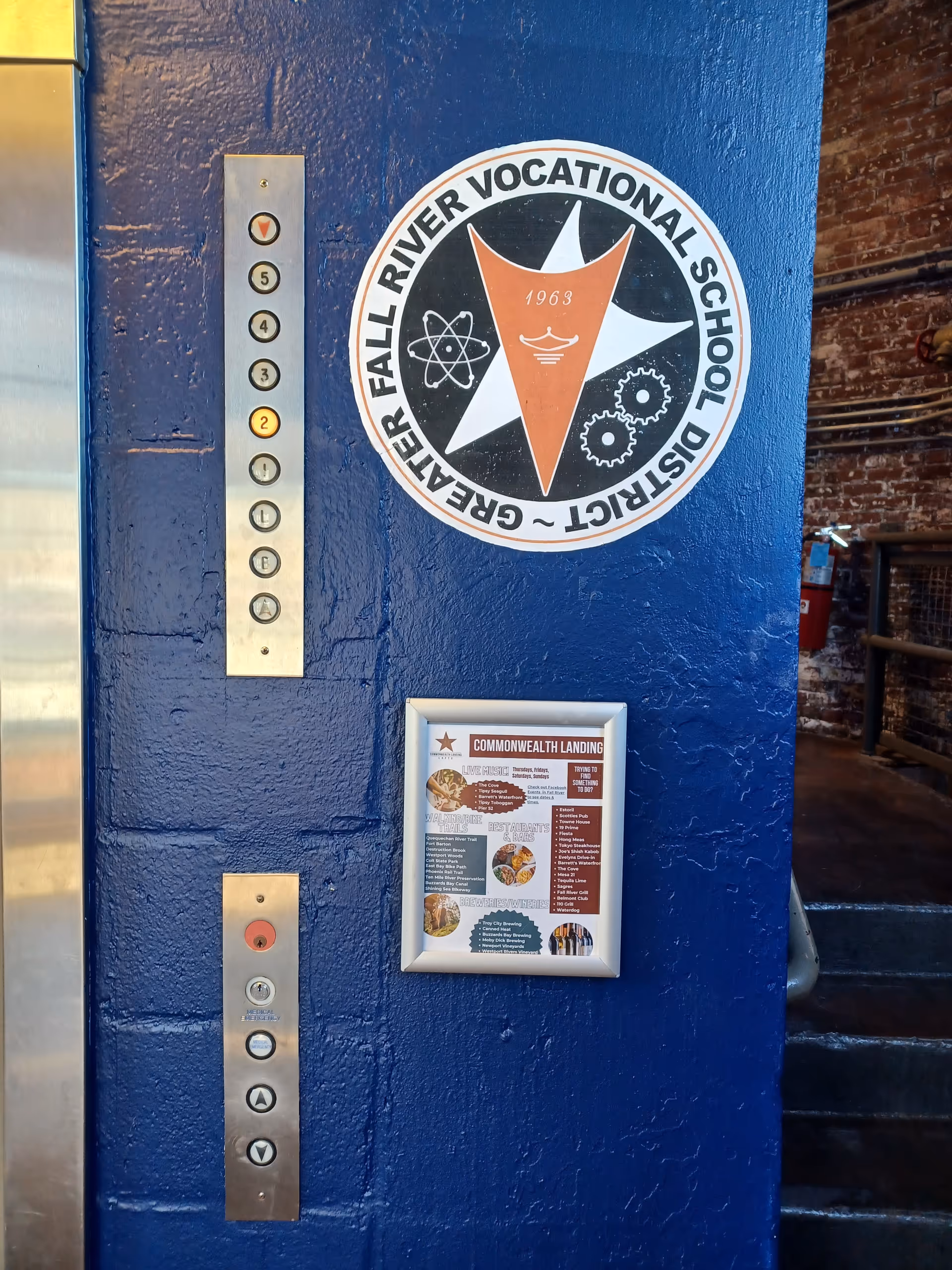 Blue wall with elevator panel, a large Fall River vocational emblem, and a framed poster.