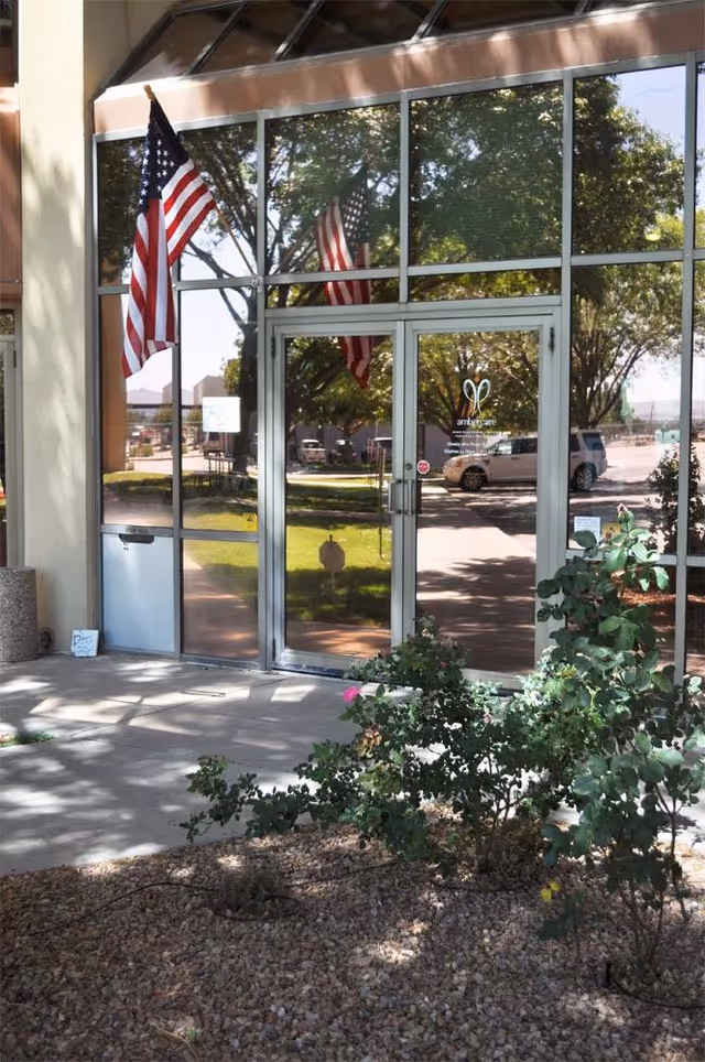 Glass entrance of Ambercare facility with American flags and landscaping outside.