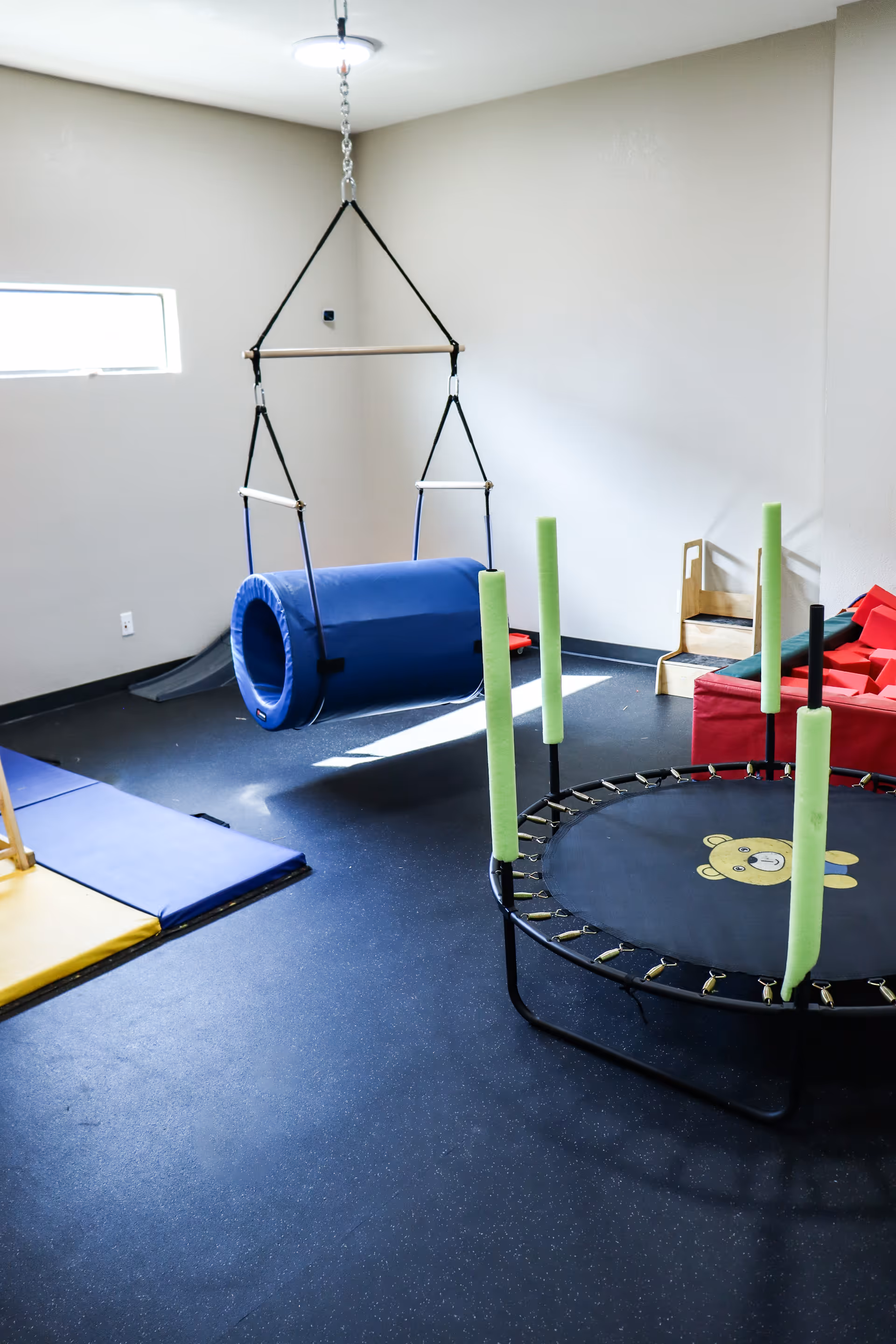 Bright therapy room with a trampoline, padded mats, and a suspended play swing.