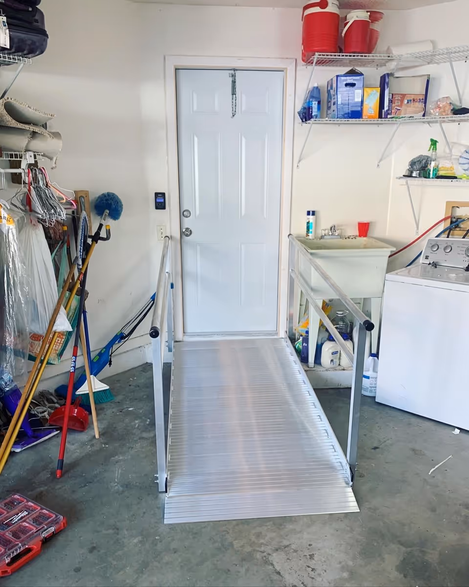 Metal wheelchair ramp leads to a white door inside a cluttered utility room with cleaning supplies.