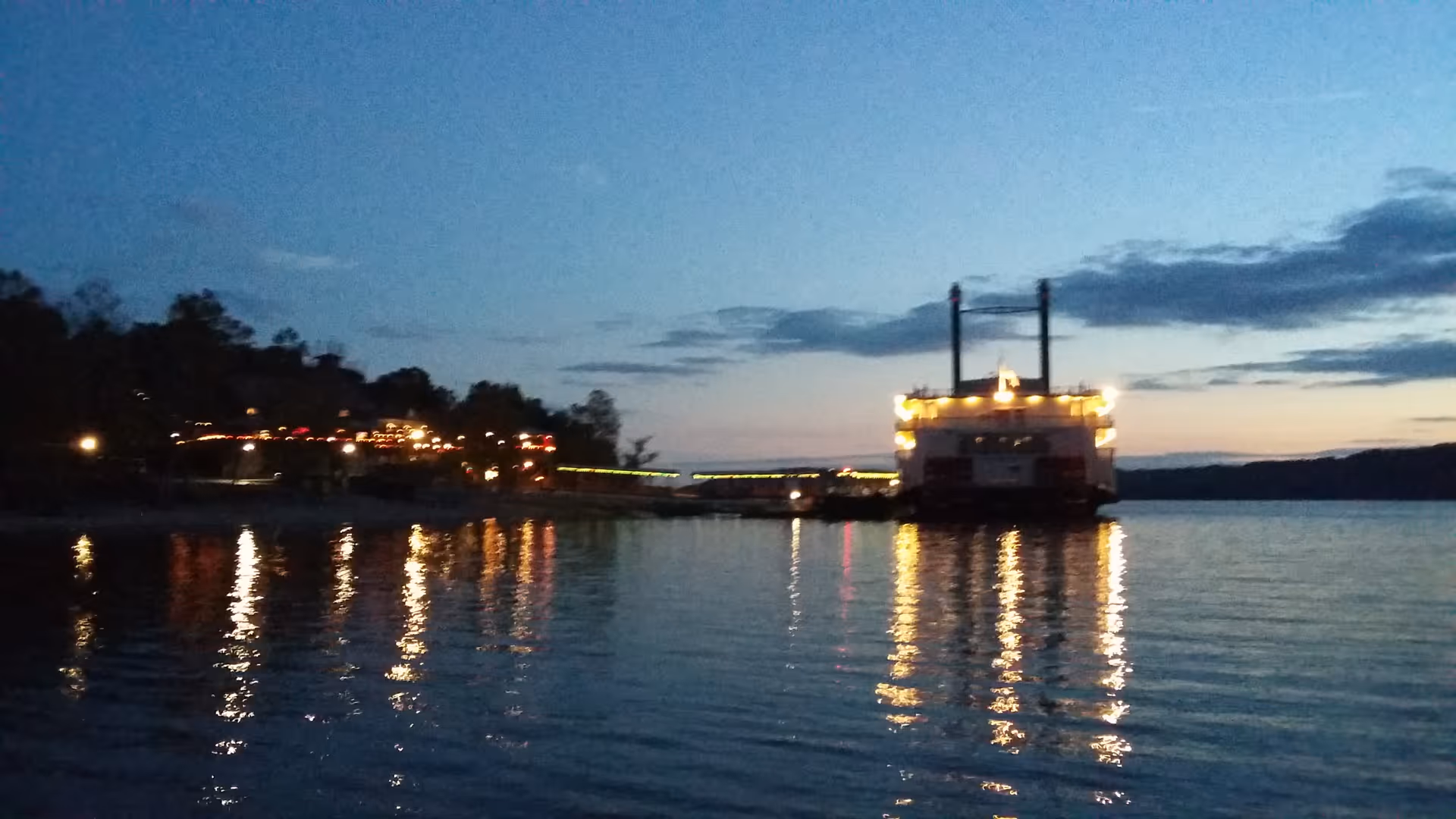 Evening waterfront scene with a lit boat and shoreline lights reflecting on water.