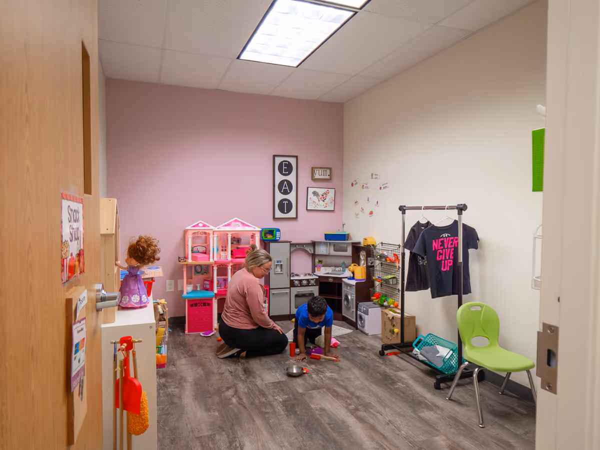 Adult caregiver and child play in a small toy-filled therapy room.