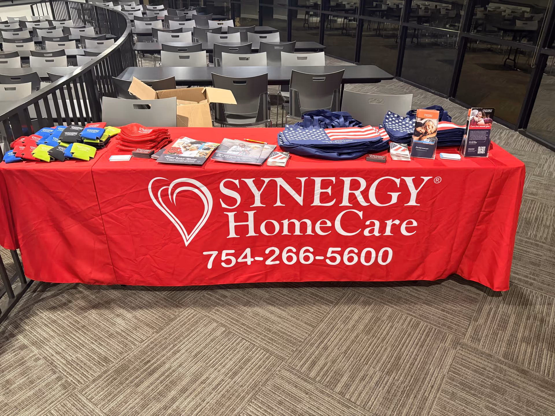 Promo table with SYNERGY HomeCare branding and branded merchandise inside a conference area.