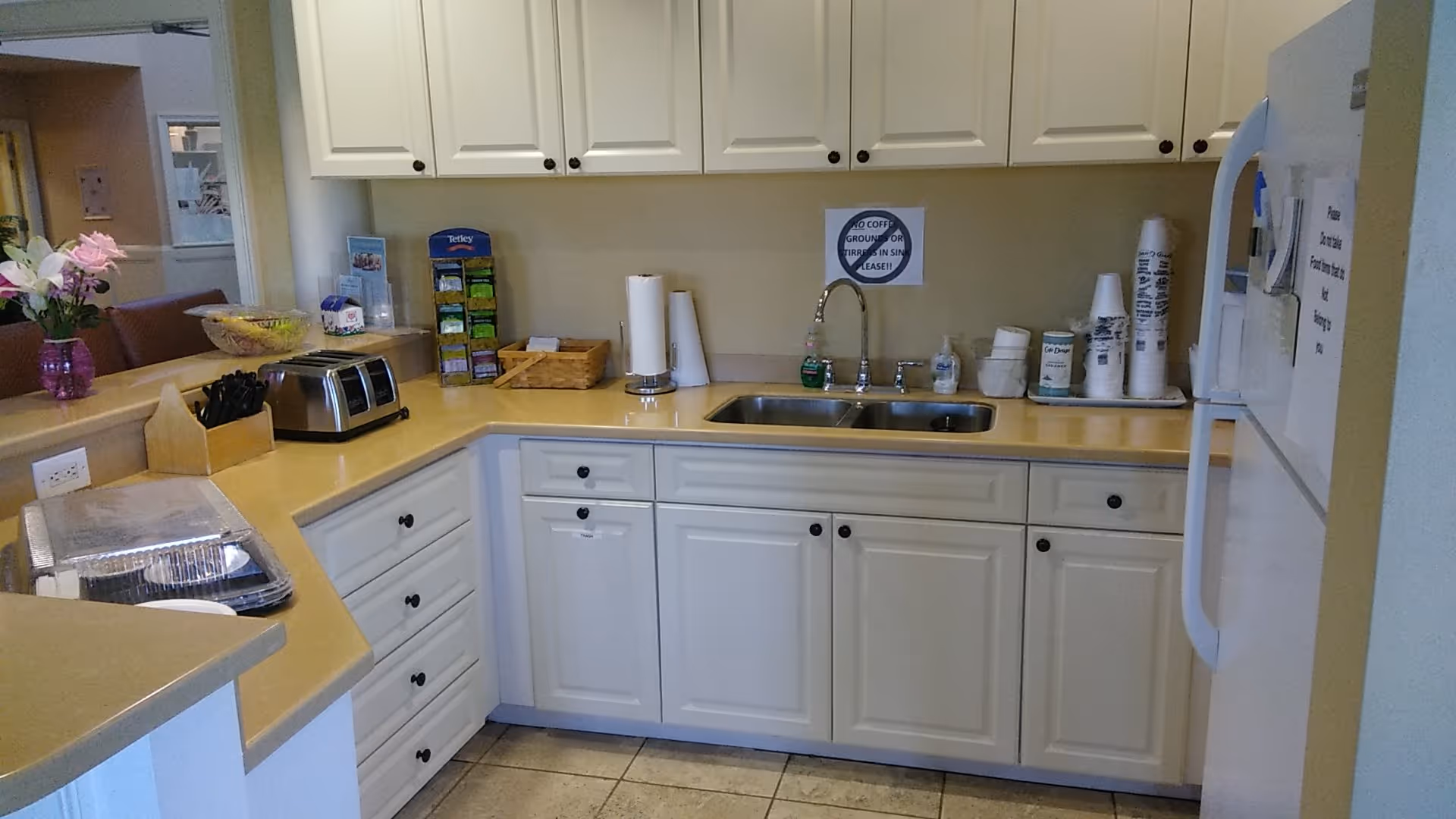 Bright white kitchenette with sink, cabinets, toaster, coffee station, and flowers on counter.