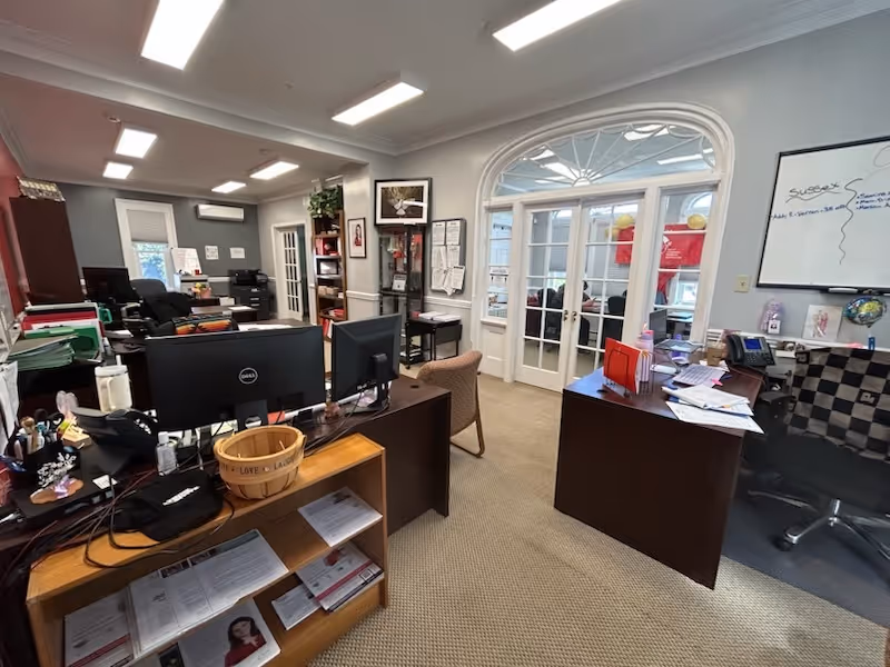 Office workspace with desks, computers, filing shelves, and glass double doors in a bright facility.