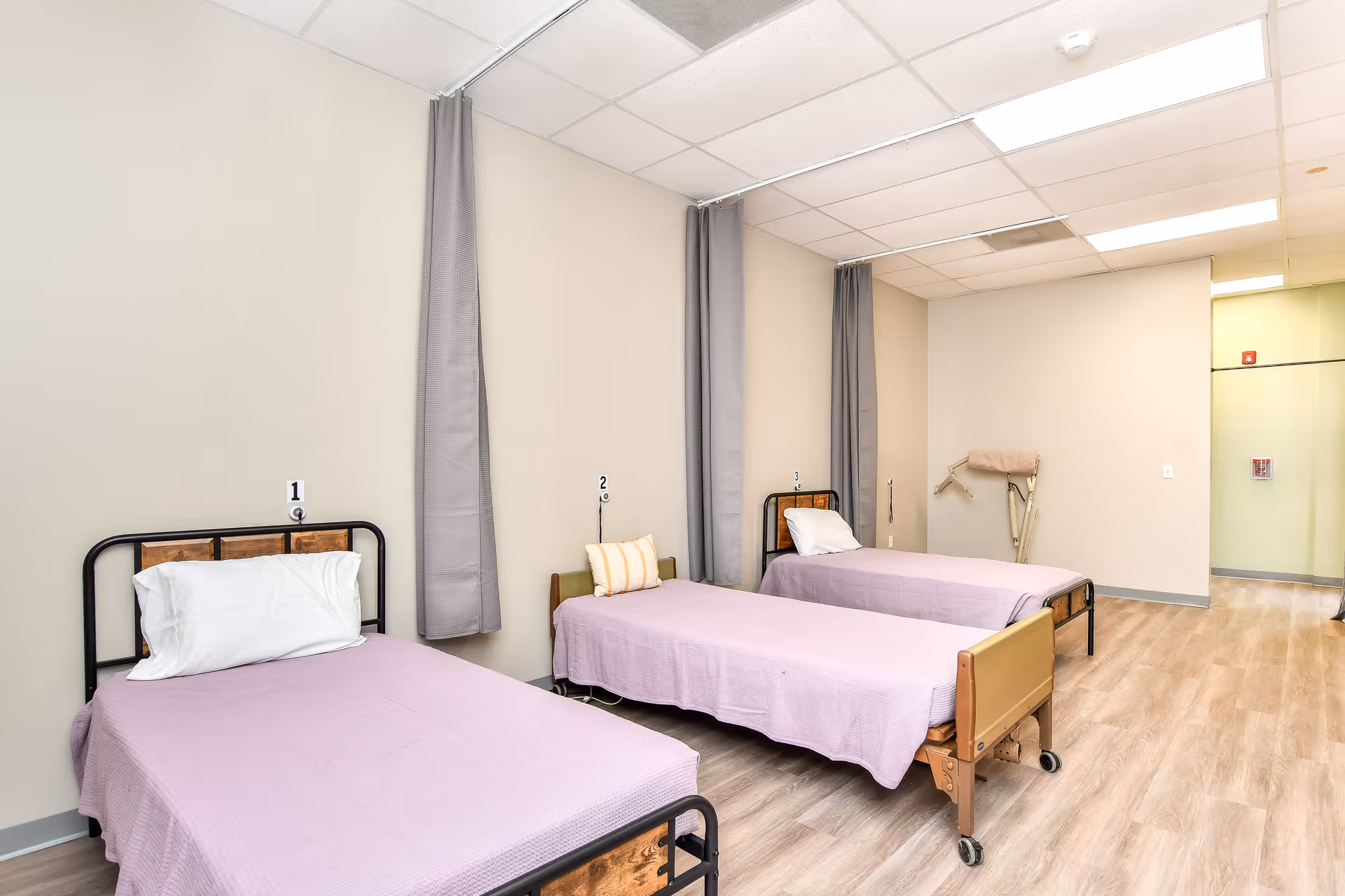 Three hospital-style beds with lavender linens and privacy curtains in a shared senior living bedroom.