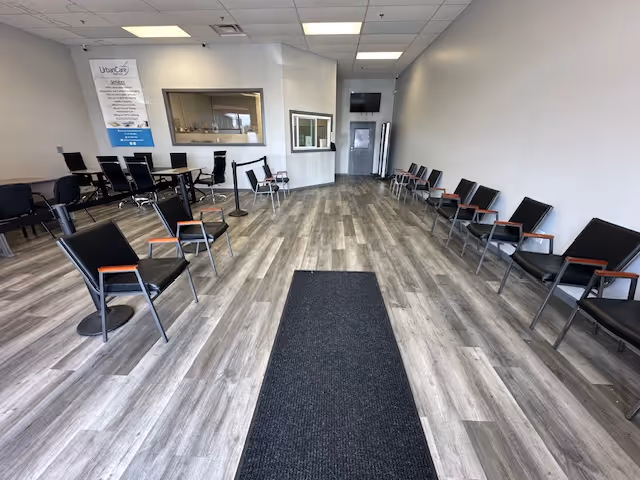 Wide waiting area with rows of black chairs along walls, central carpet, and reception window.