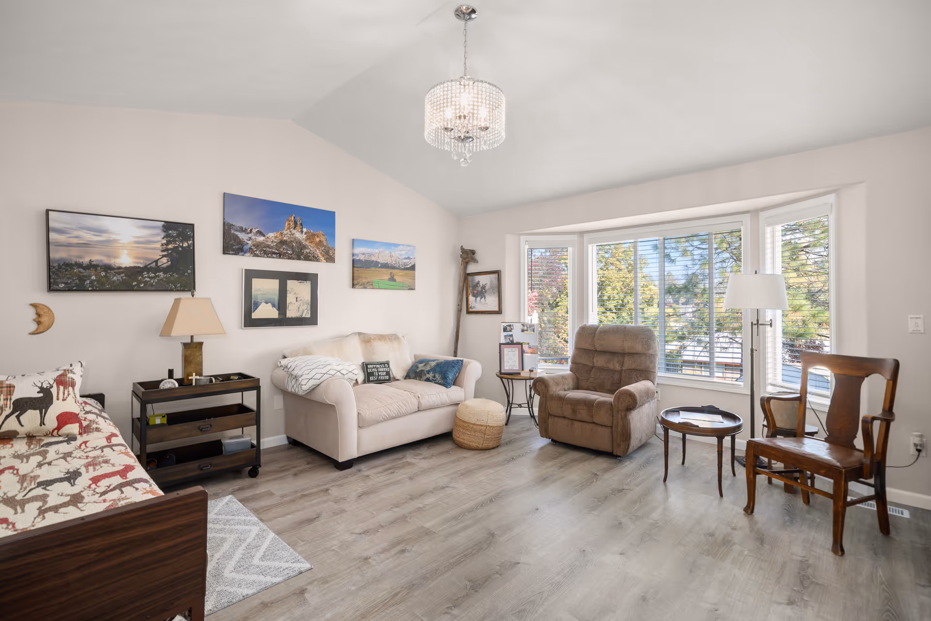 Bright living room with beige sofa, recliner, wooden chair, bay window, and wall art.