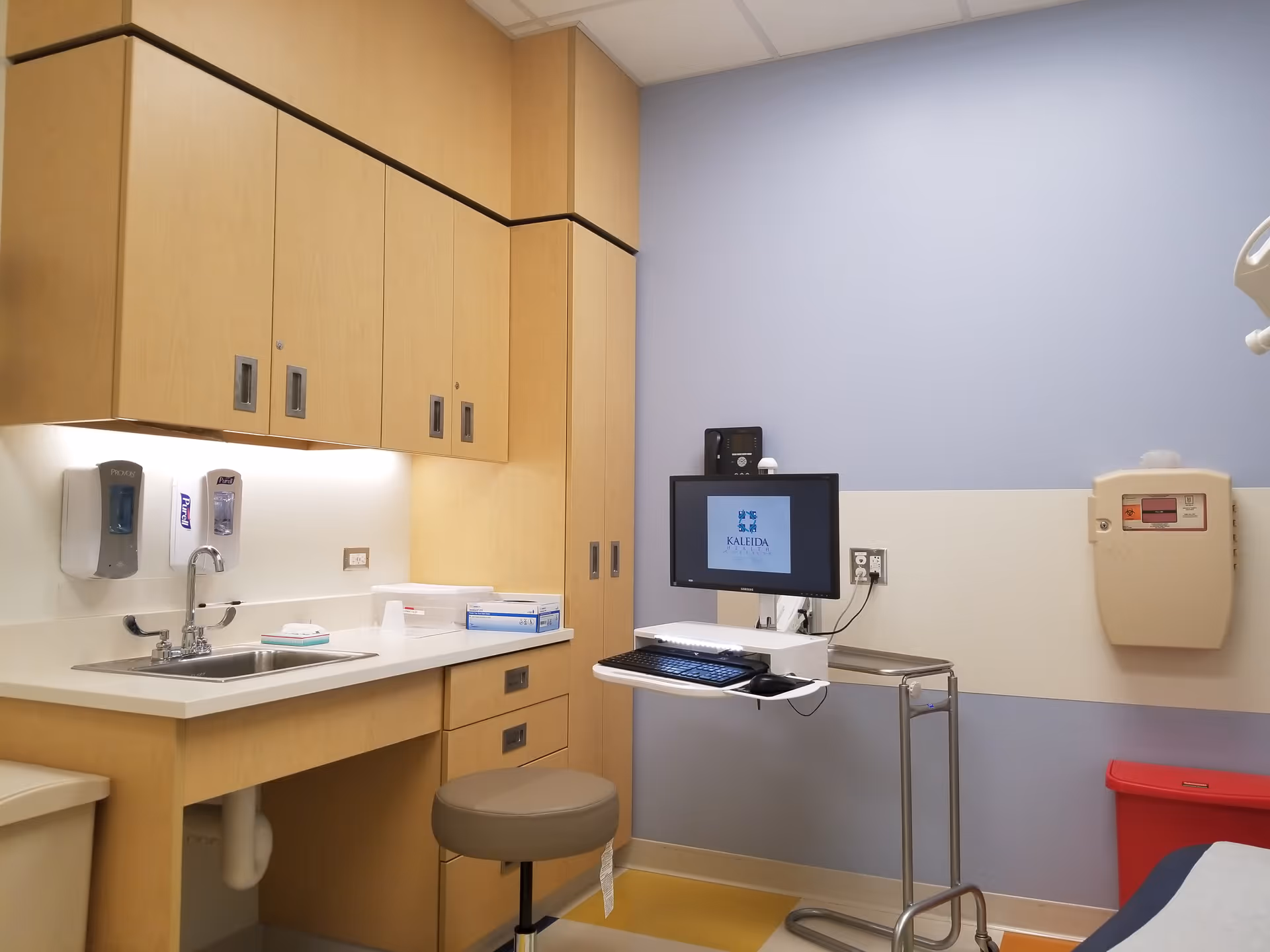 A medical examination room with wooden cabinets, a sink, computer station, and medical supplies.