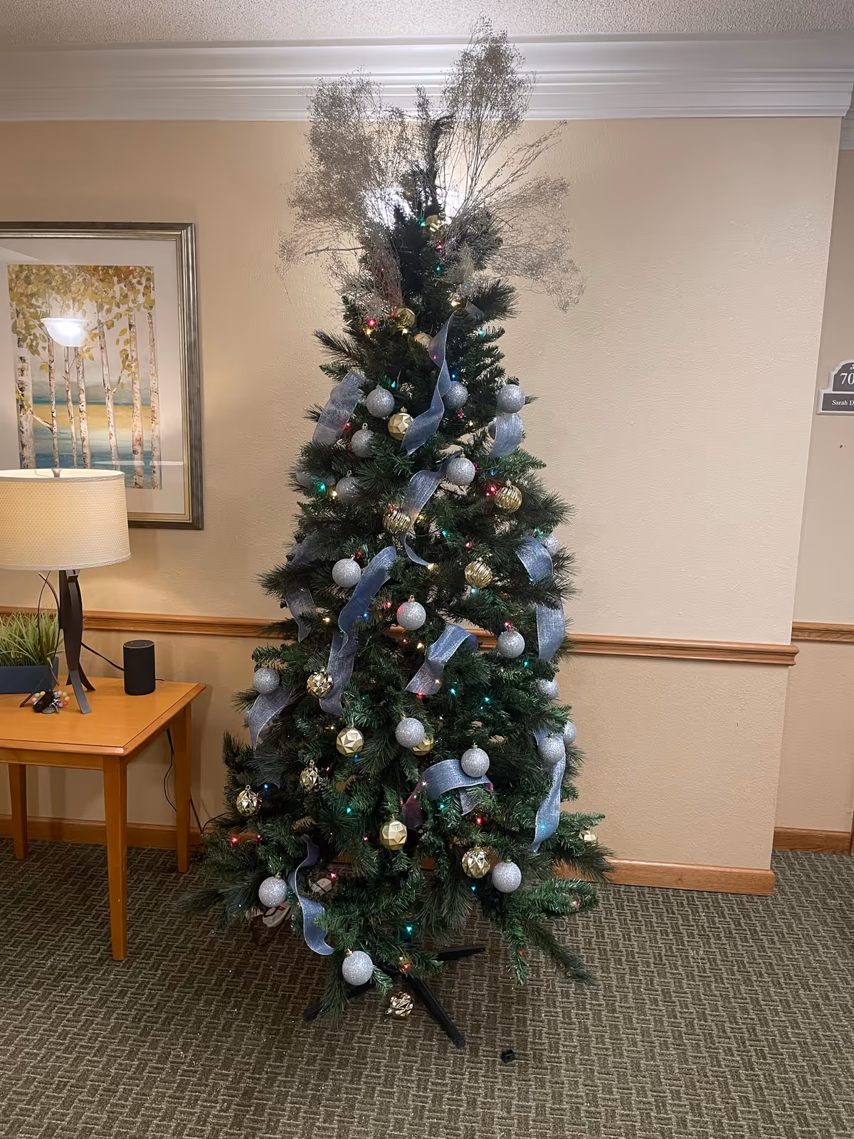 Decorated Christmas tree with silver ornaments stands in a hallway beside a table and lamp.