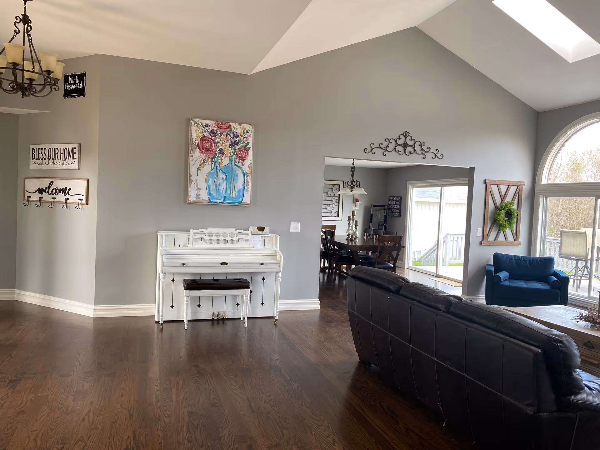 Bright living room with dark leather sofa, white piano, and arched window overlooking outdoors.