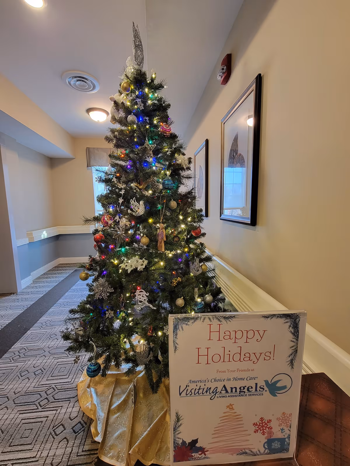 Decorated Christmas tree in a hallway with a holiday sign from Visiting Angels.