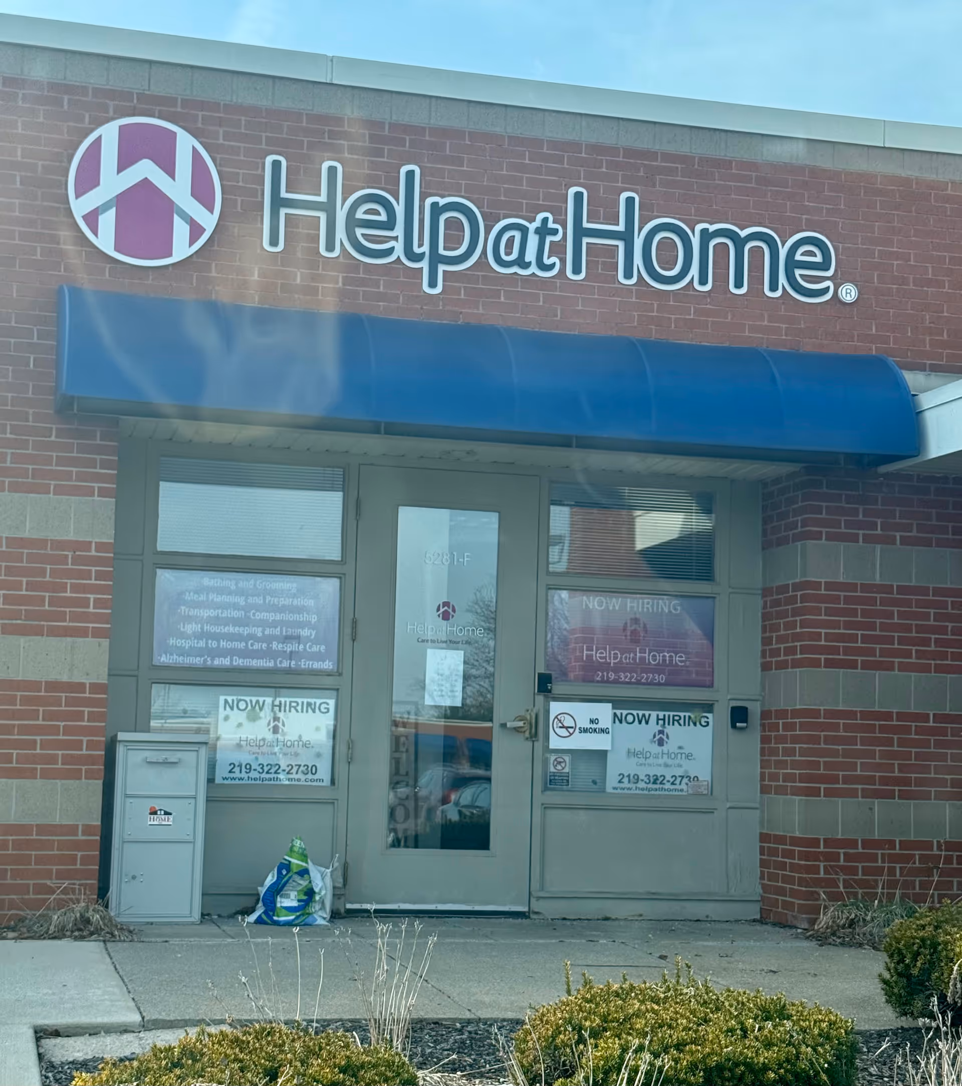 Brick building entrance with Help at Home sign, blue awning, glass doors, and now hiring posters.