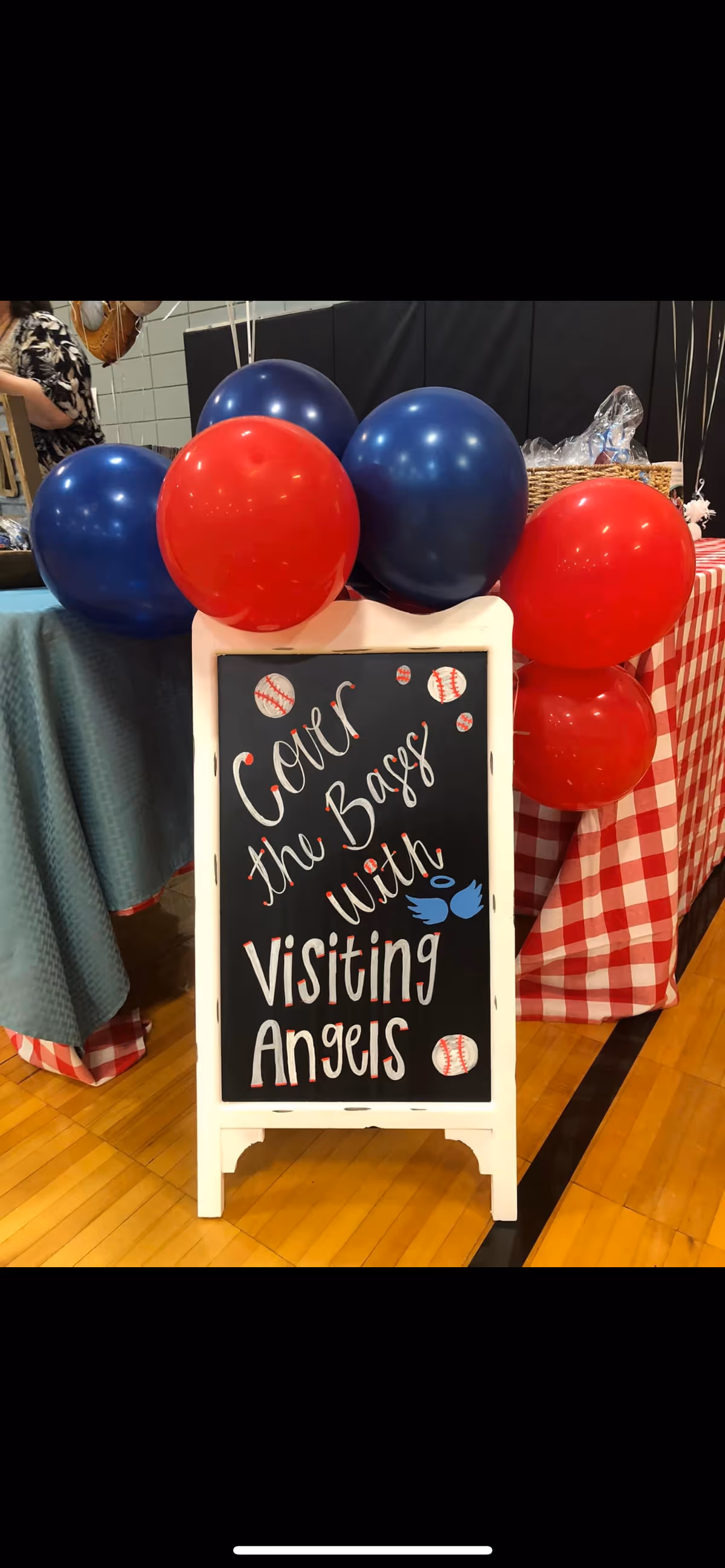 Indoor scene with a chalkboard sign reading Visiting Angels, flanked by red and blue balloons.
