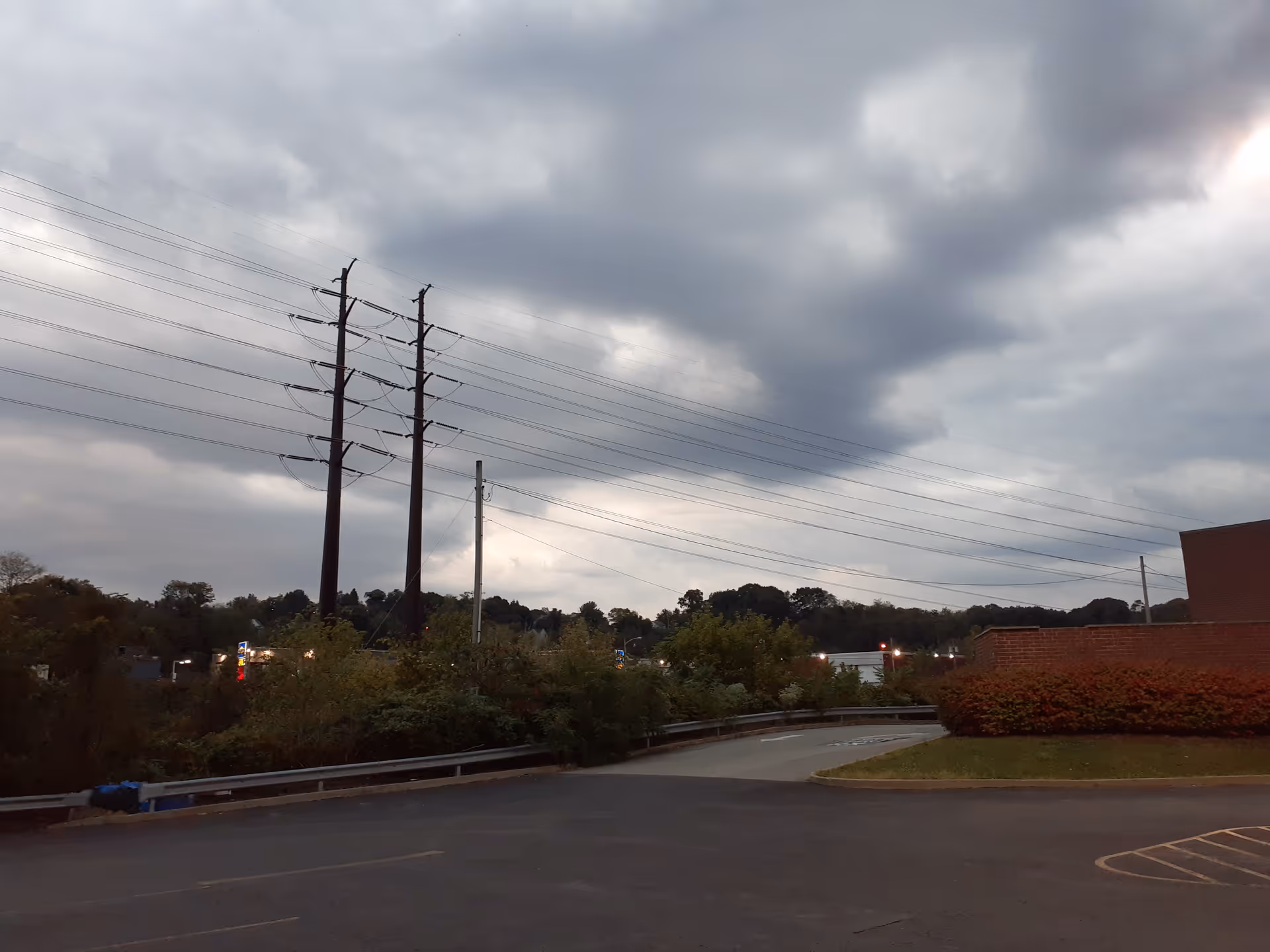 Overcast sky above a parking lot with utility poles and shrubbery along a road.