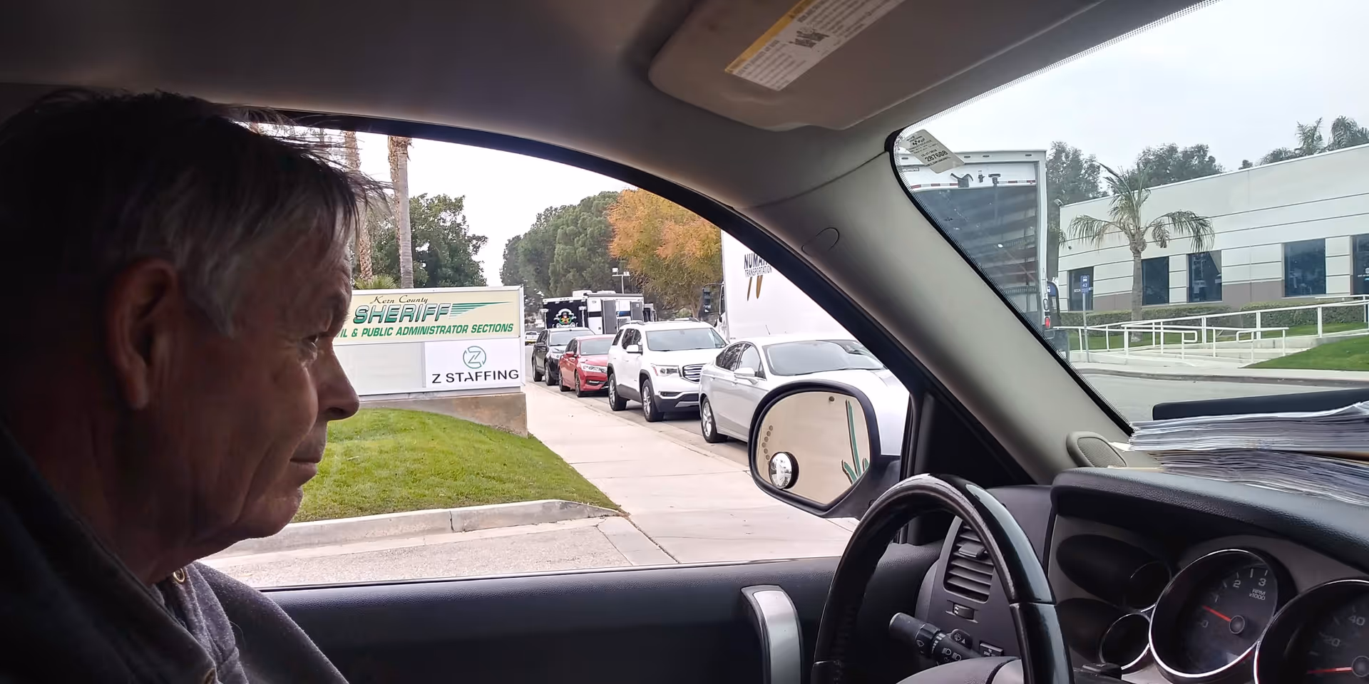 An elderly man sits in a car, gazing at a building and parked vehicles outside.