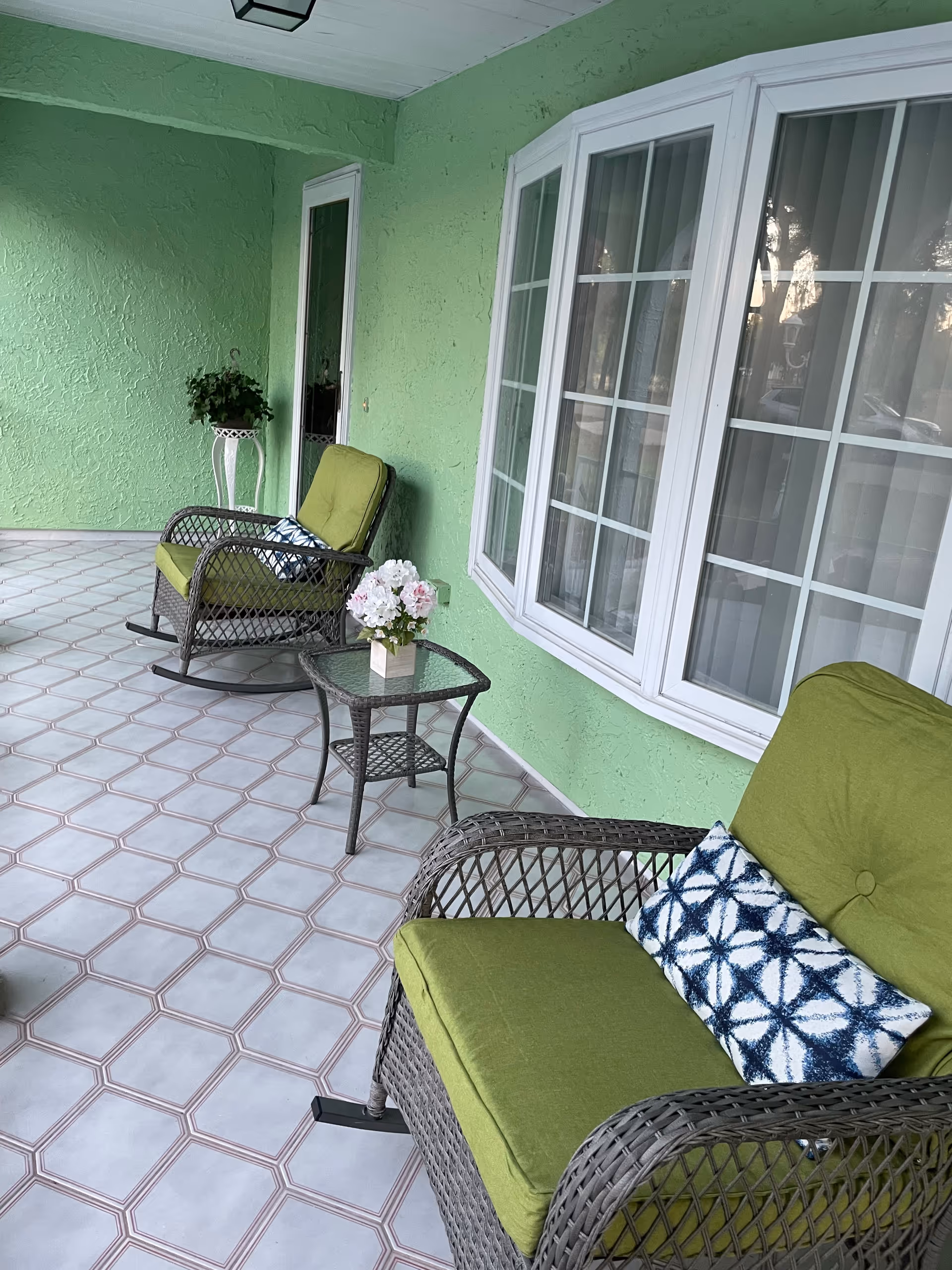 Green exterior porch with wicker seating, glass table, and potted flowers.