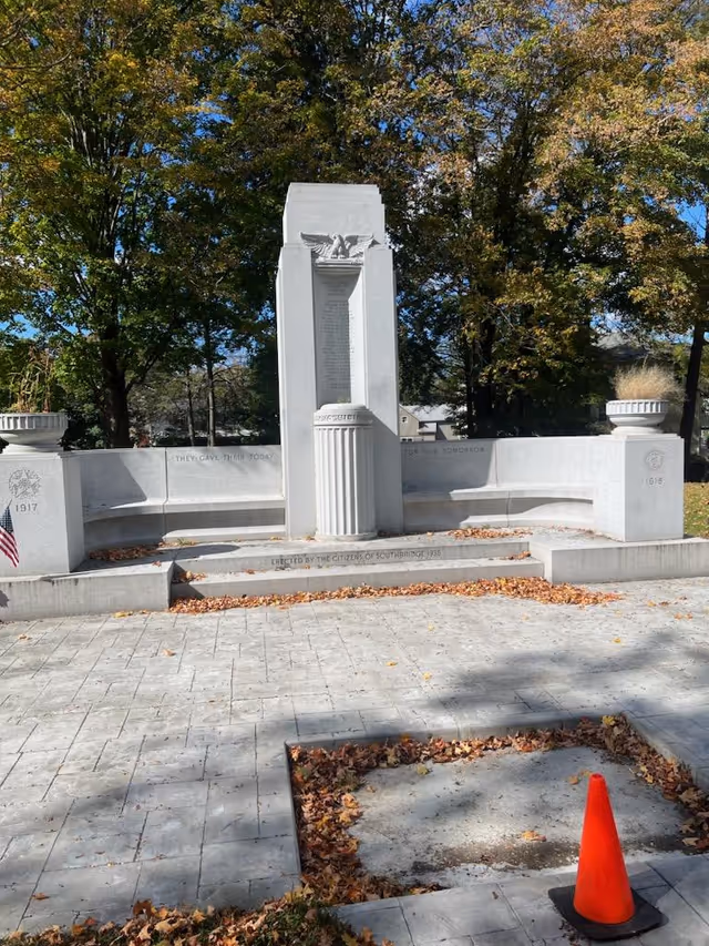 Outdoor memorial monument with stone benches and leaf-covered ground, trees in background.