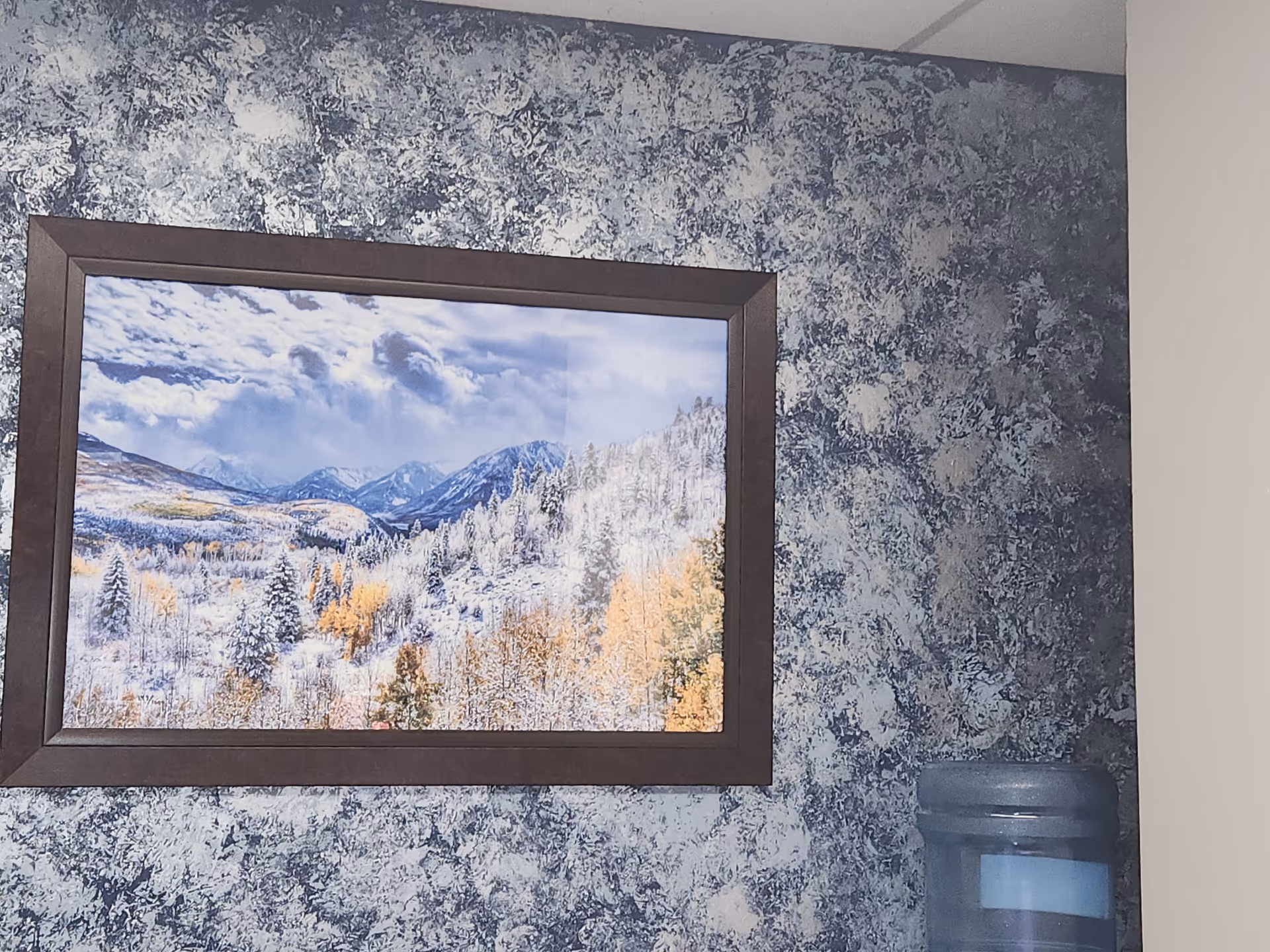 Framed landscape photo on a blue speckled wall beside a water cooler.