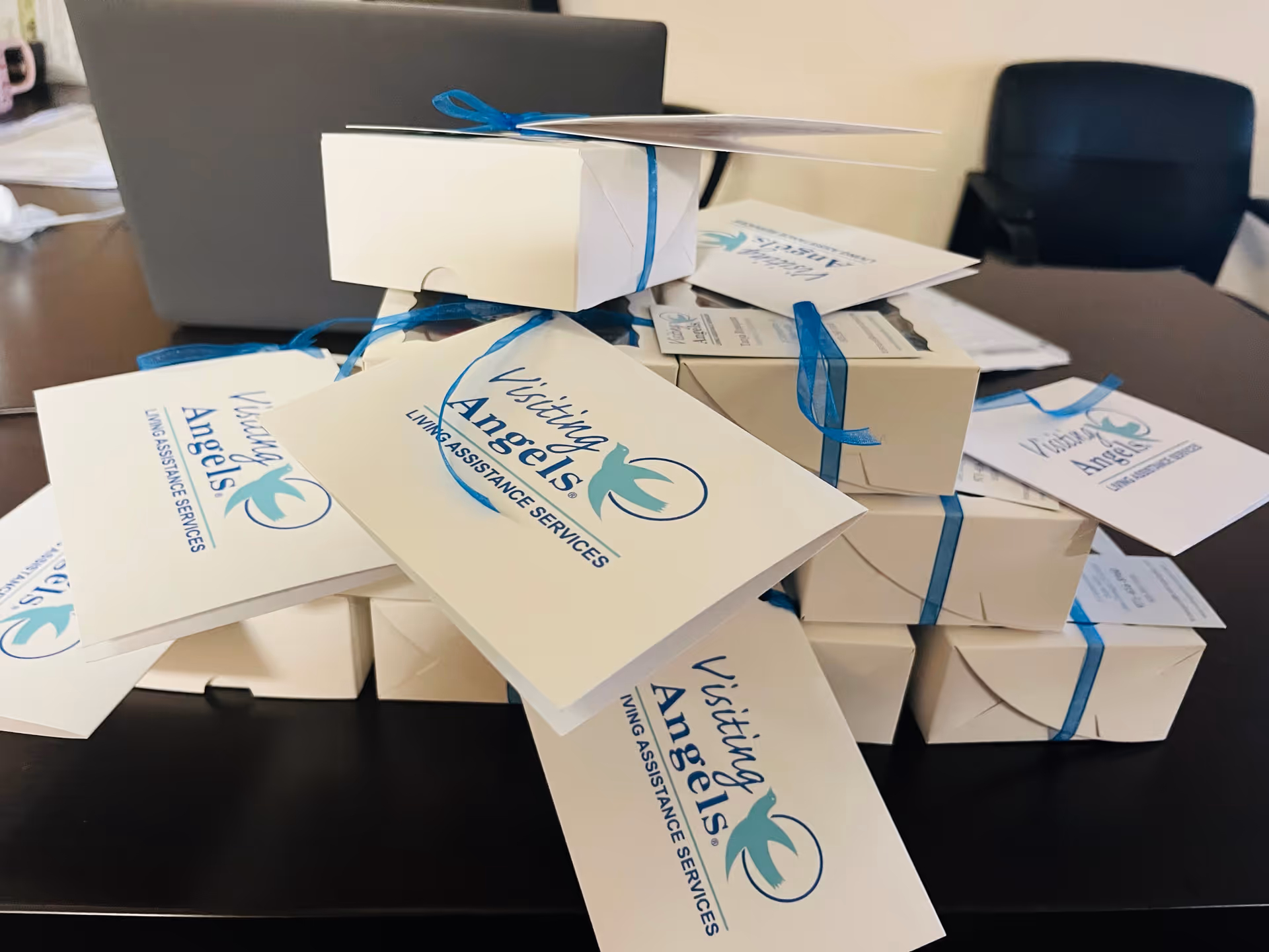 Desk cluttered with white gift boxes tied with blue ribbons and Visiting Angels cards.