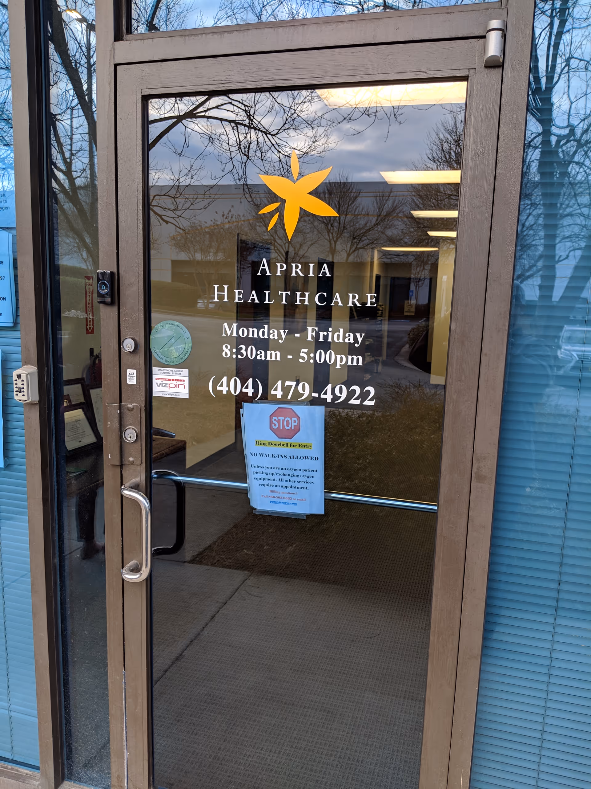 Glass entrance door with Apria Healthcare branding and hours, reflections of trees outside