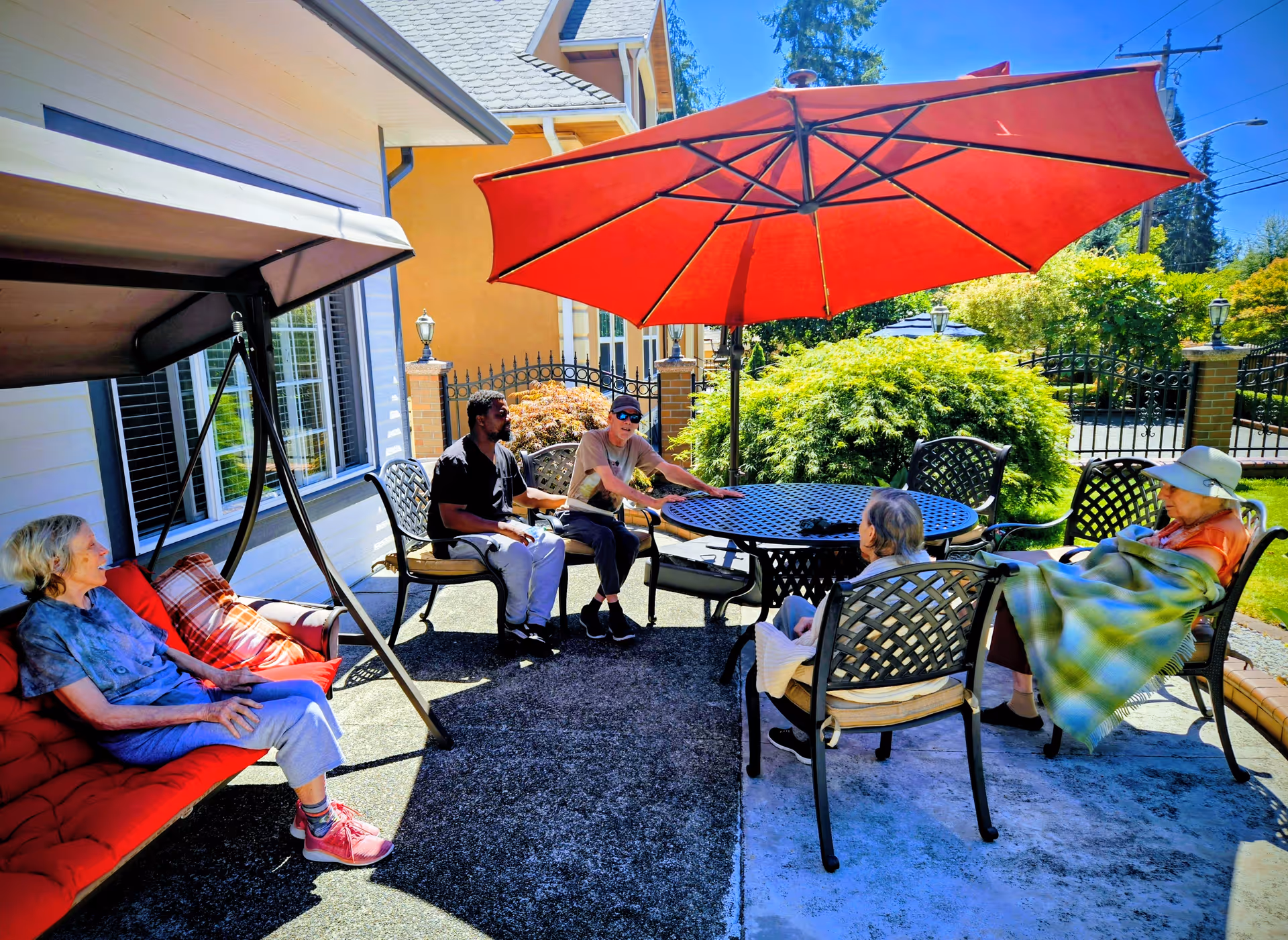 Group of seniors sits and chats around an outdoor patio table under a large red umbrella.
