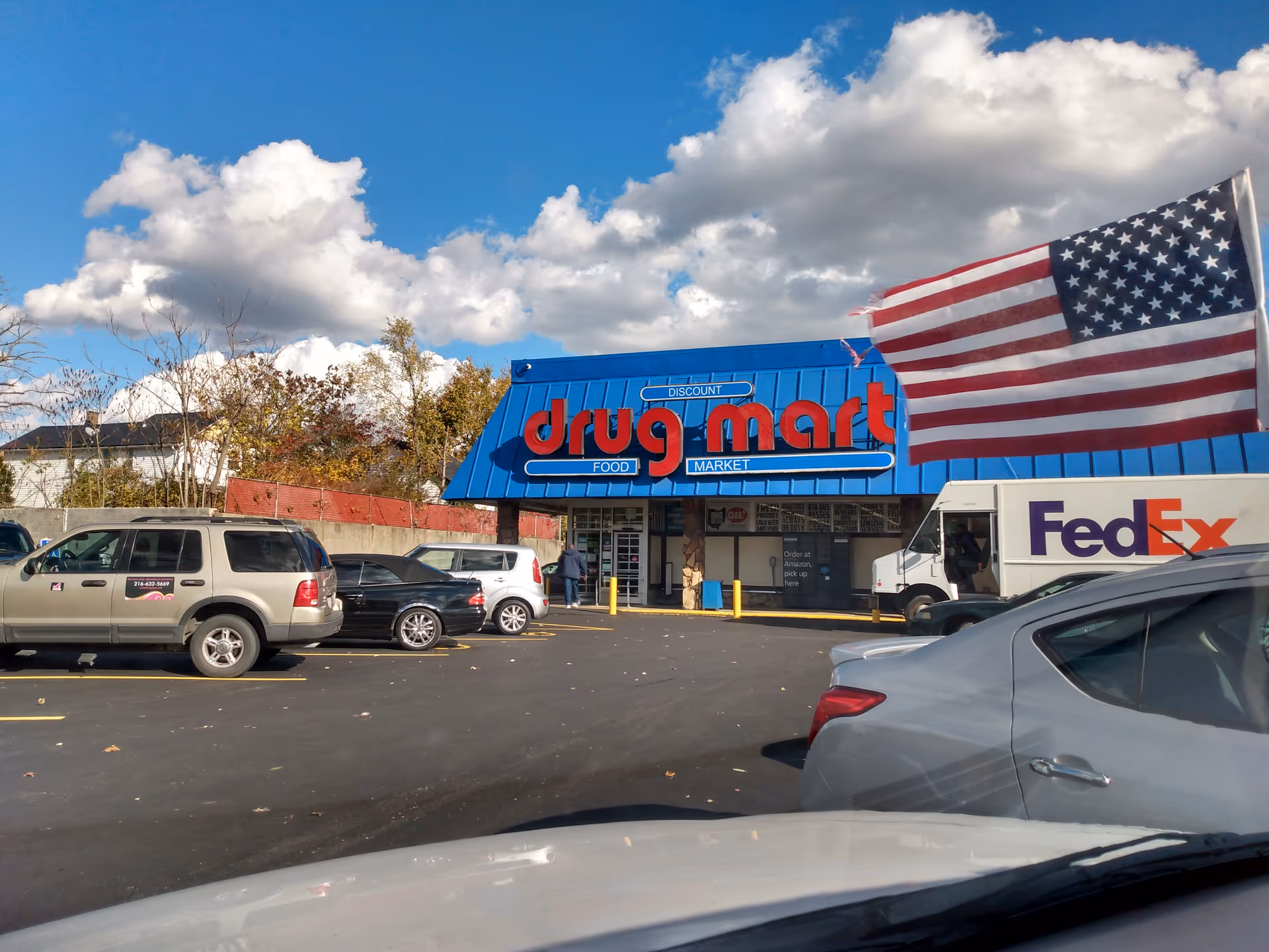 Blue Discount Drug Mart storefront with American flag, parked cars, and a FedEx truck.