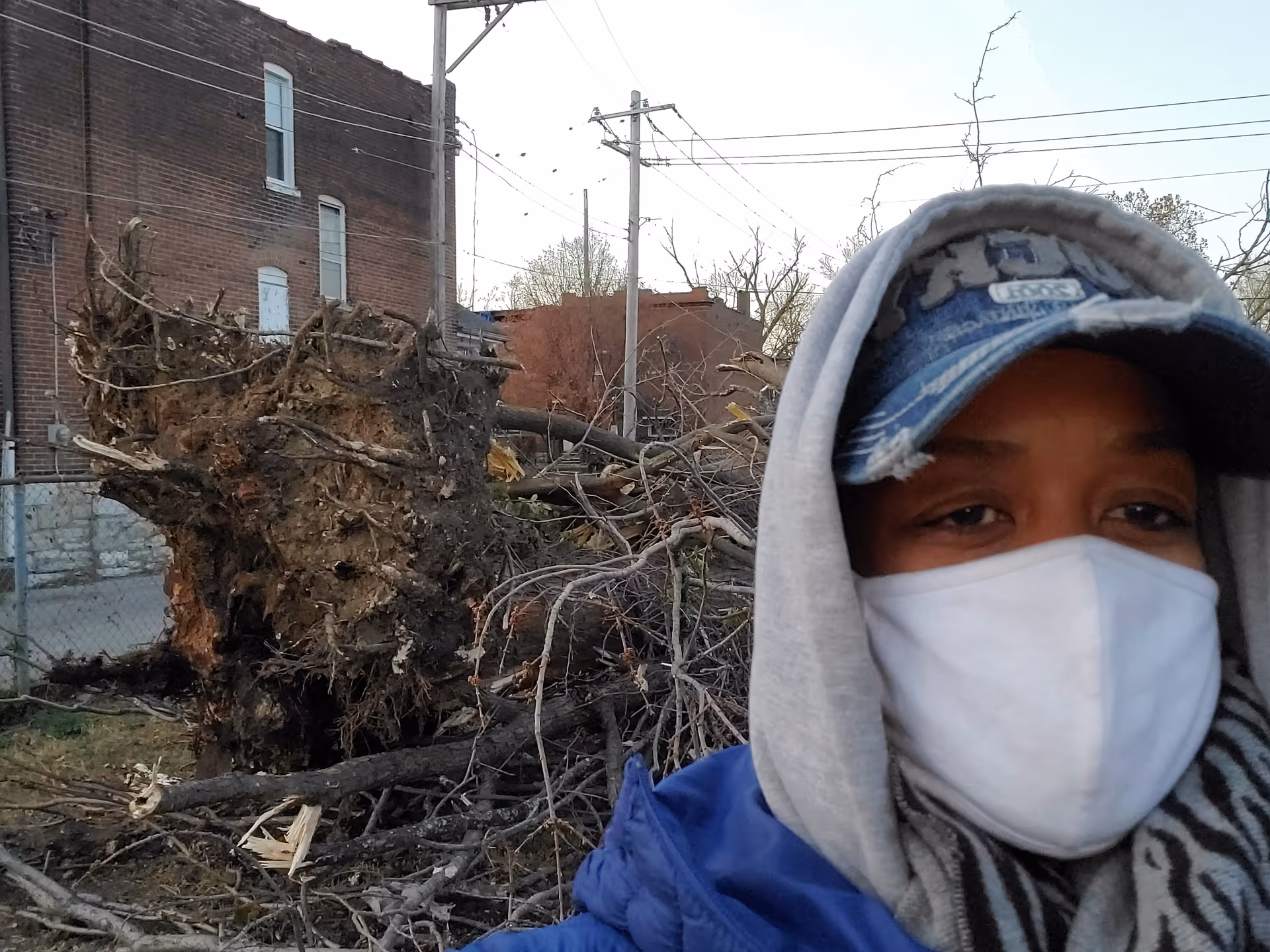 Person wearing a mask and hoodie stands beside uprooted tree debris outside a brick building.