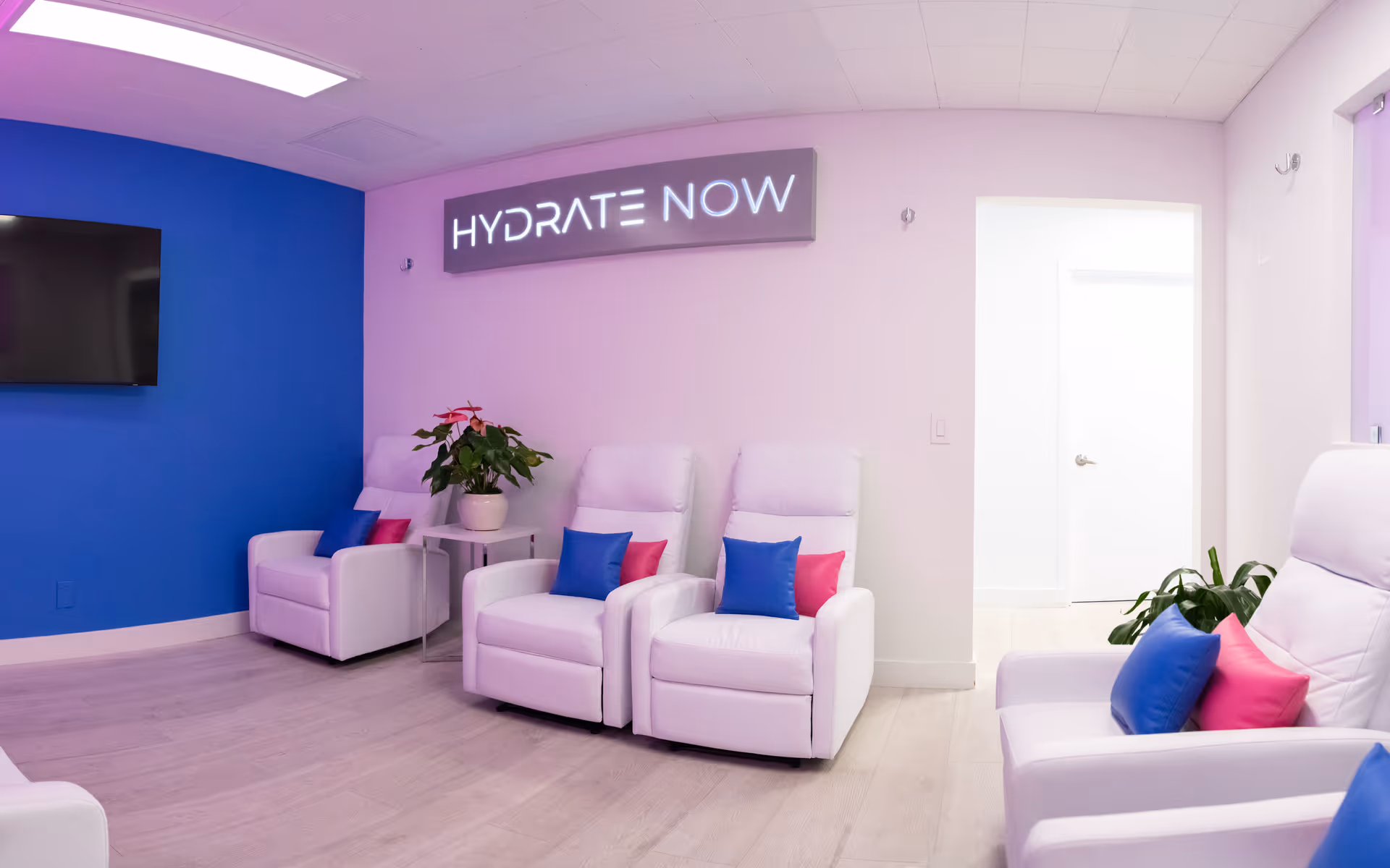 A pastel-lit lounge area with white recliners, blue and pink pillows, and a Hydrate Now sign.