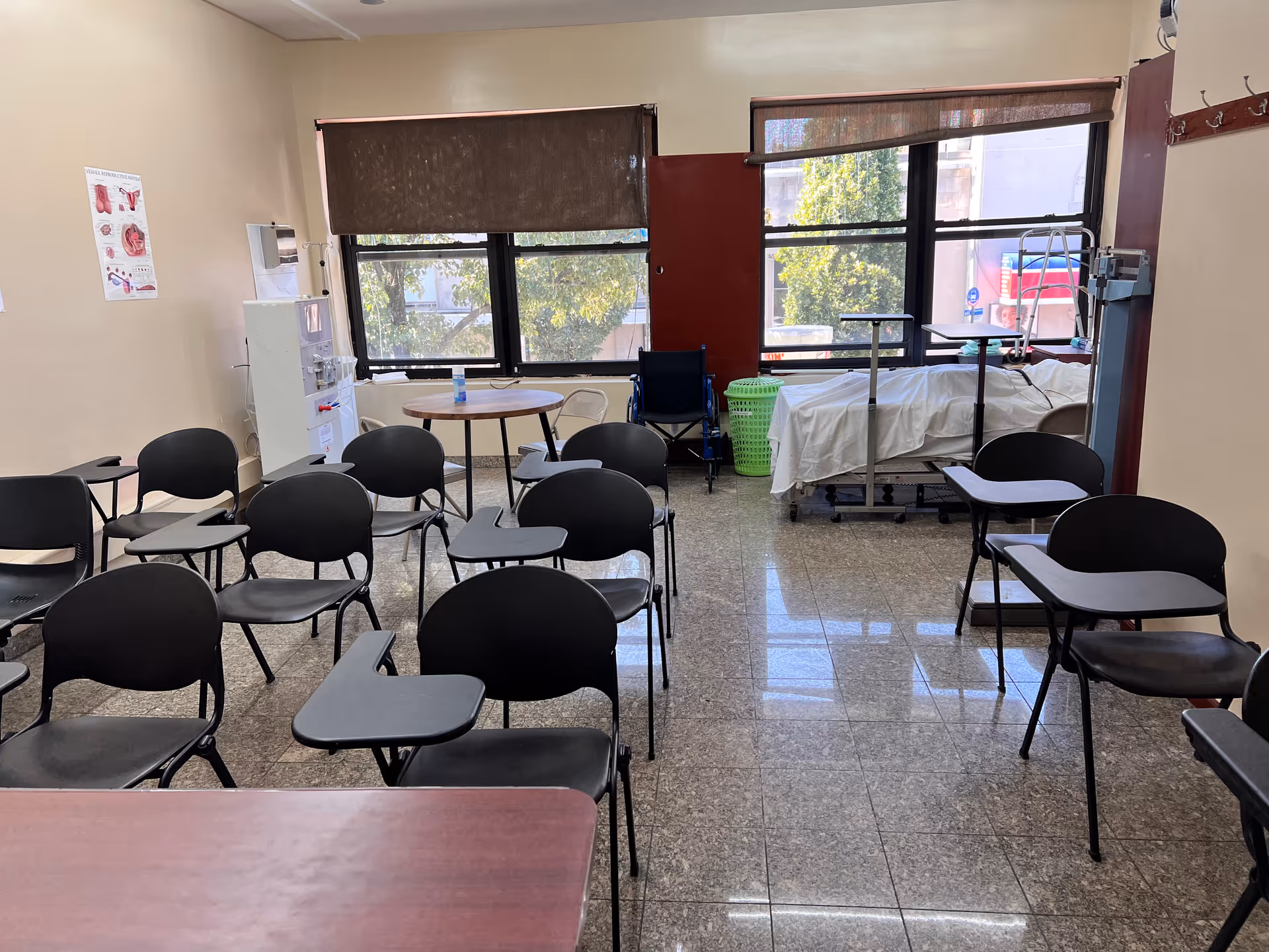 An interior classroom-style room with multiple black chairs and a hospital bed near large windows.