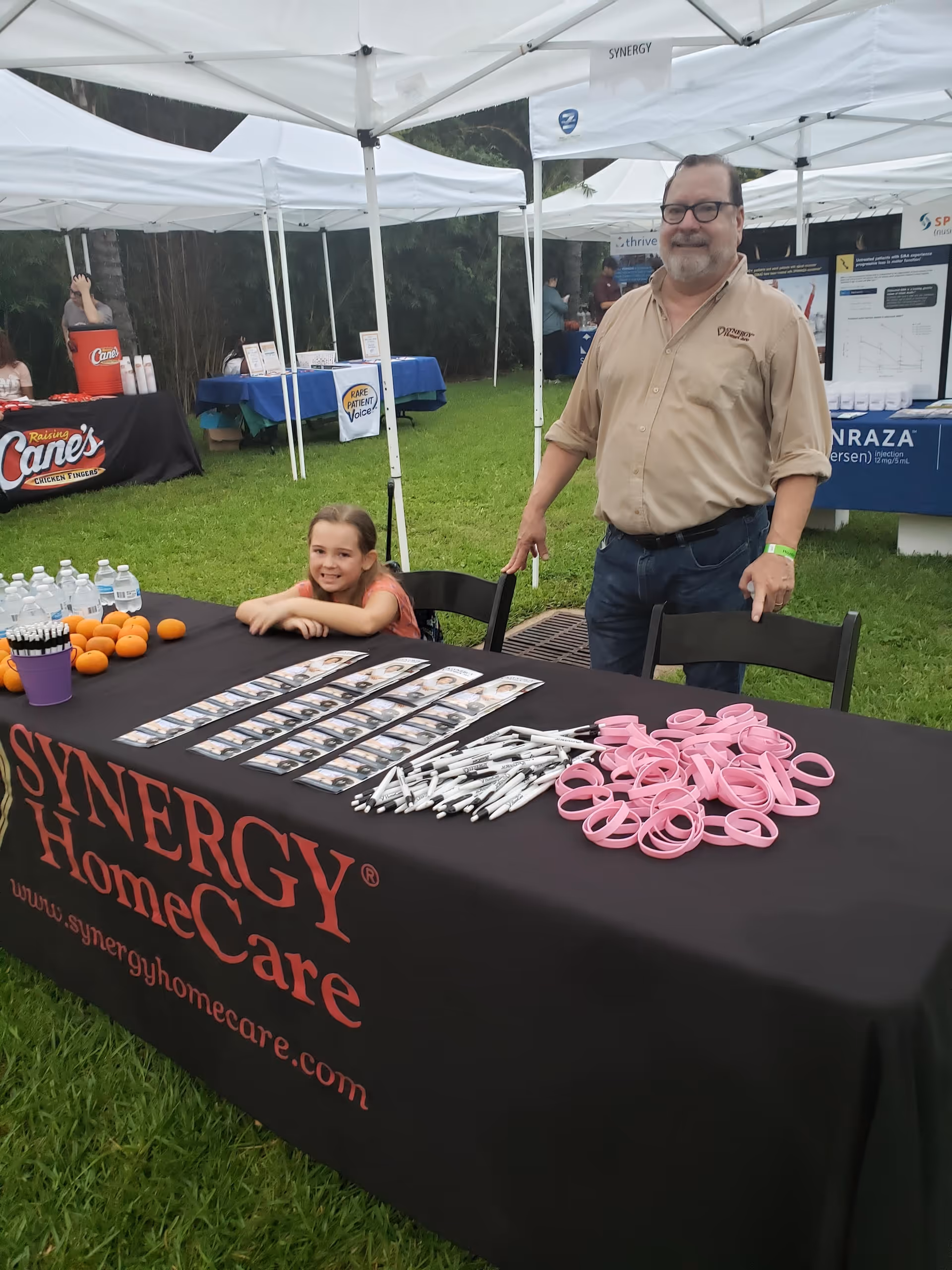A man and young girl stand beside a Synergy HomeCare outdoor booth with promotional items.