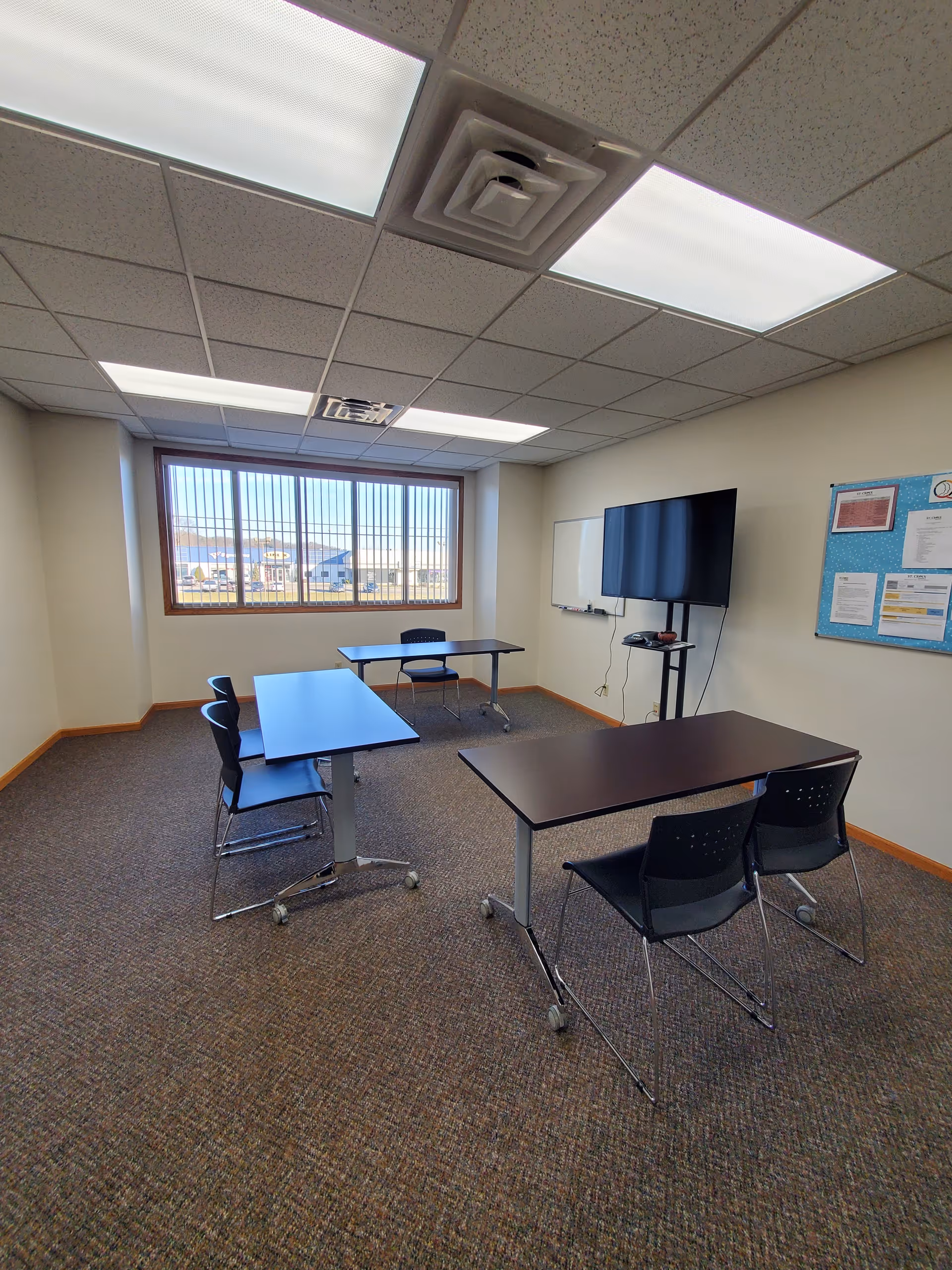 A small conference room with tables, chairs, a whiteboard, and a mounted monitor.