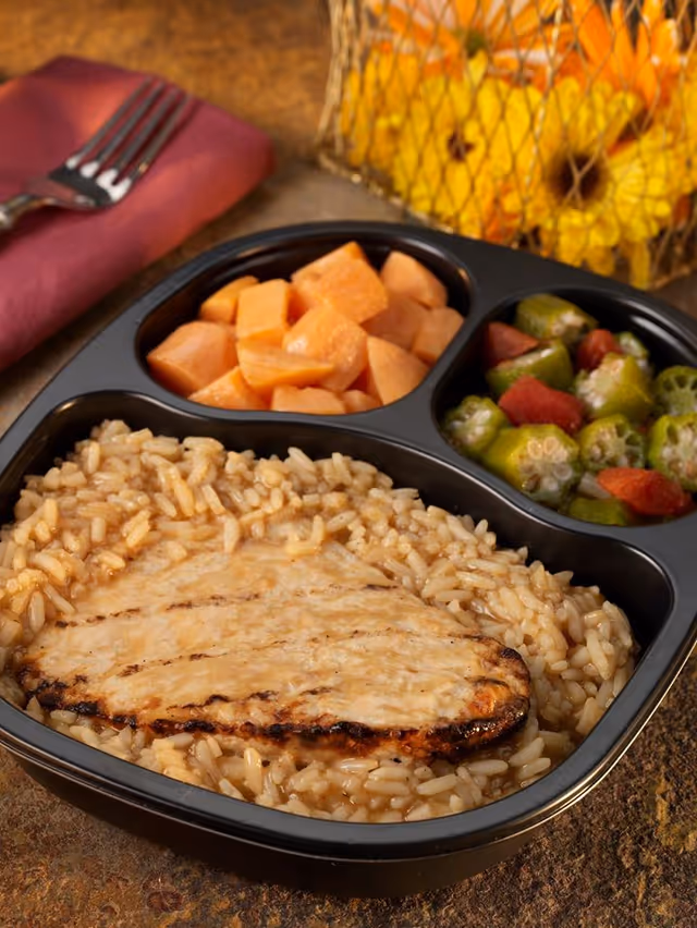 Close-up of a divided meal tray with pork, rice, and vegetables on a table.