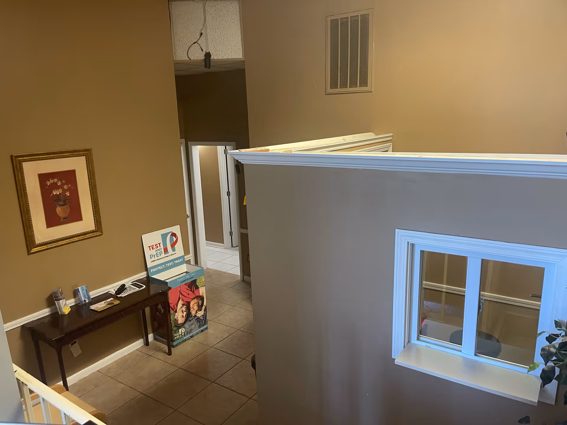 View of a beige-walled interior corner with a hallway, window, and pamphlet stand on tiled floor.