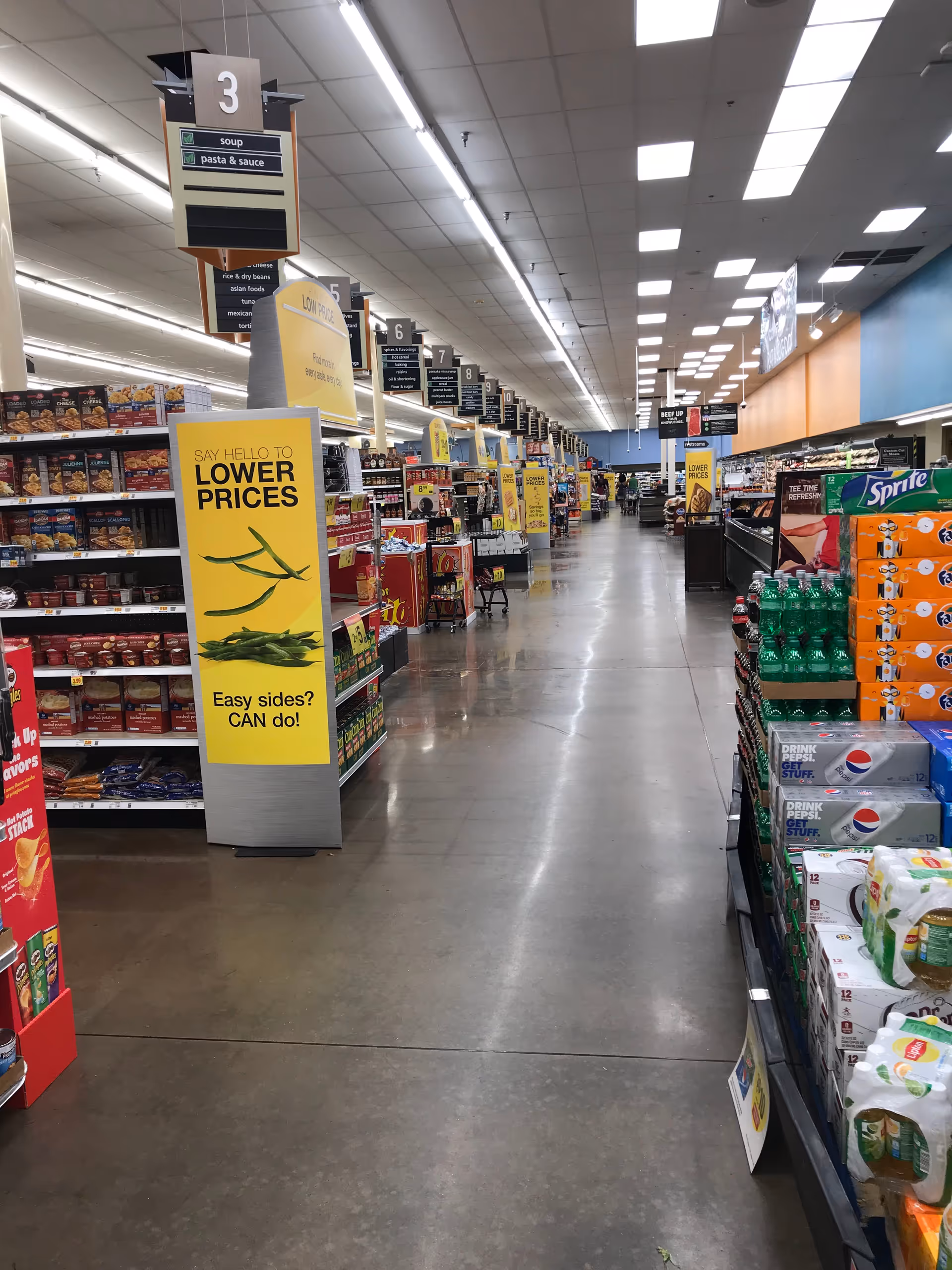 Long grocery store aisle with shelves of snacks and drinks, overhead signage.