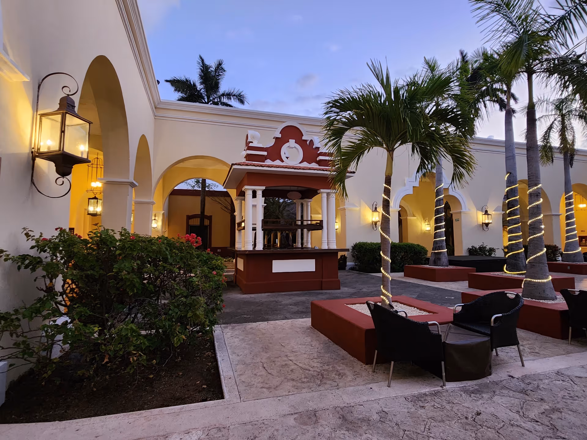 Mediterranean-style outdoor courtyard with arches, palm trees, string lights, and seating around a central kiosk.