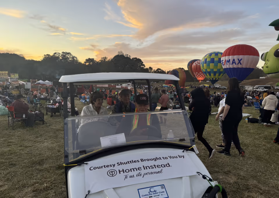 An outdoor festival scene with a golf cart banner reading Home Instead and balloons in background.