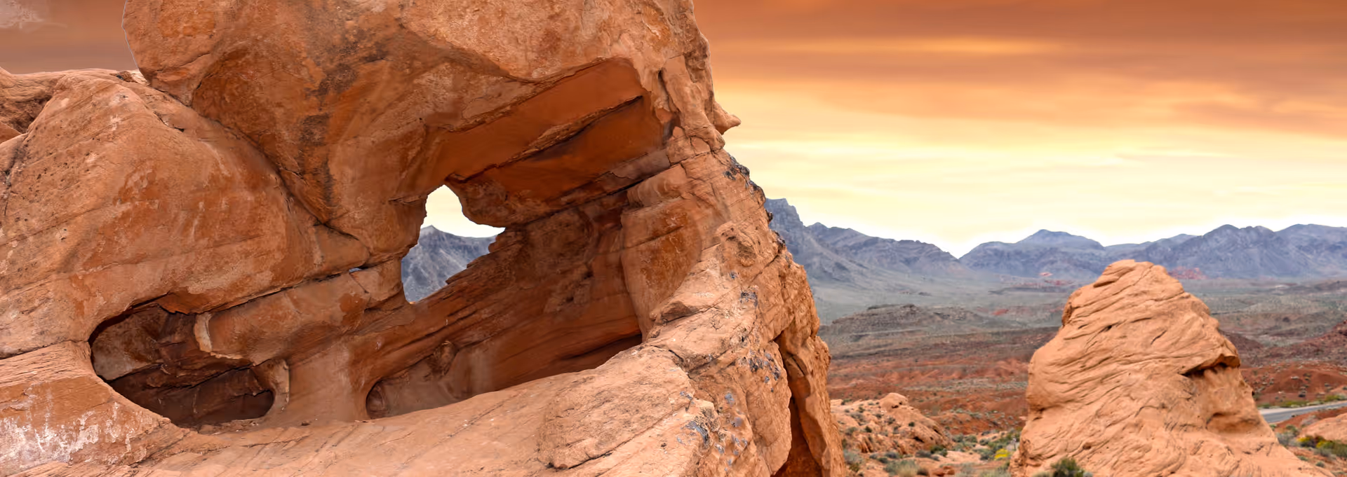 Desert landscape with a natural rock arch, distant mountains, and a sunset sky.