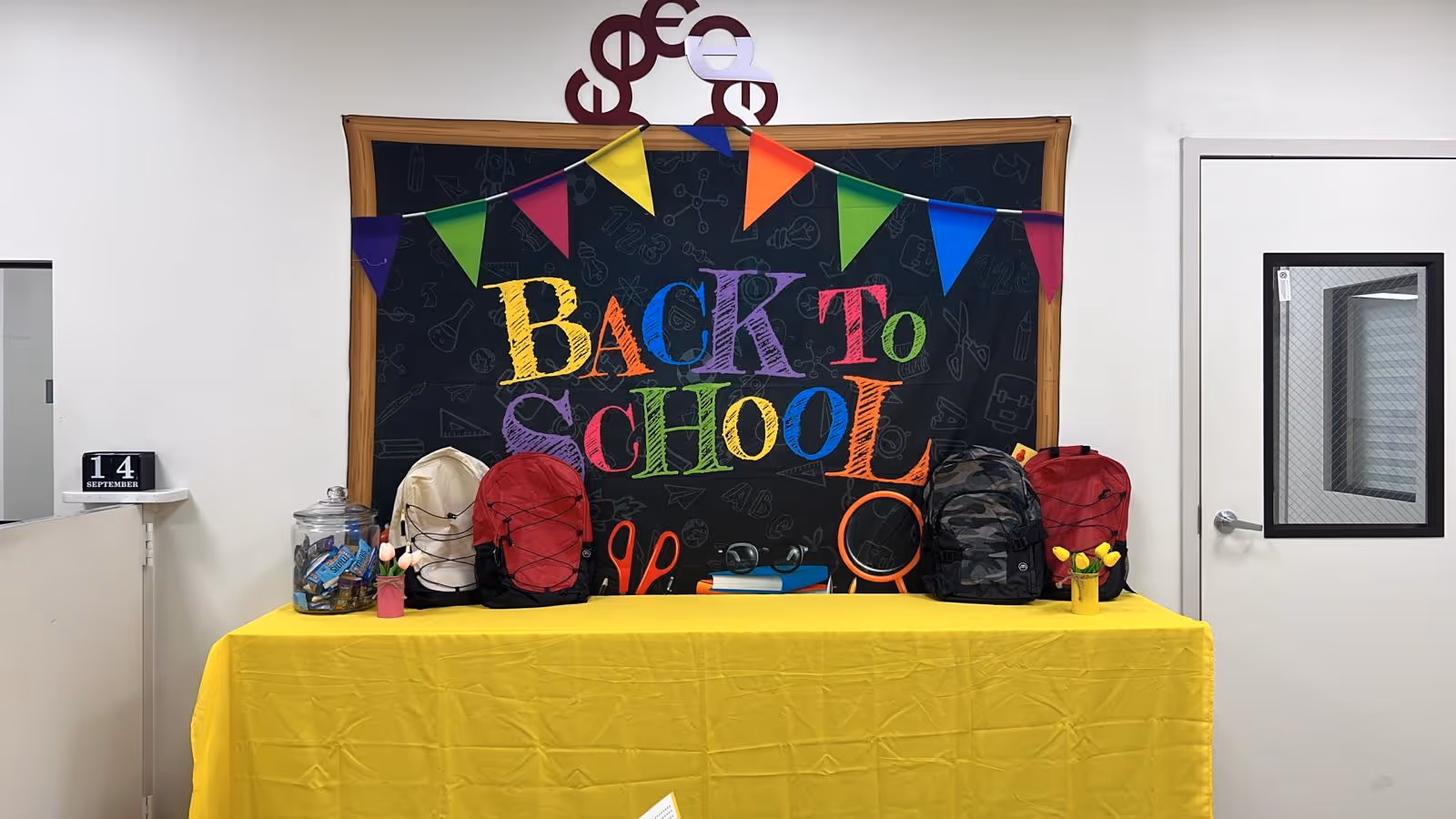 Indoor school-themed display with backpacks on a yellow table and a chalkboard backdrop.
