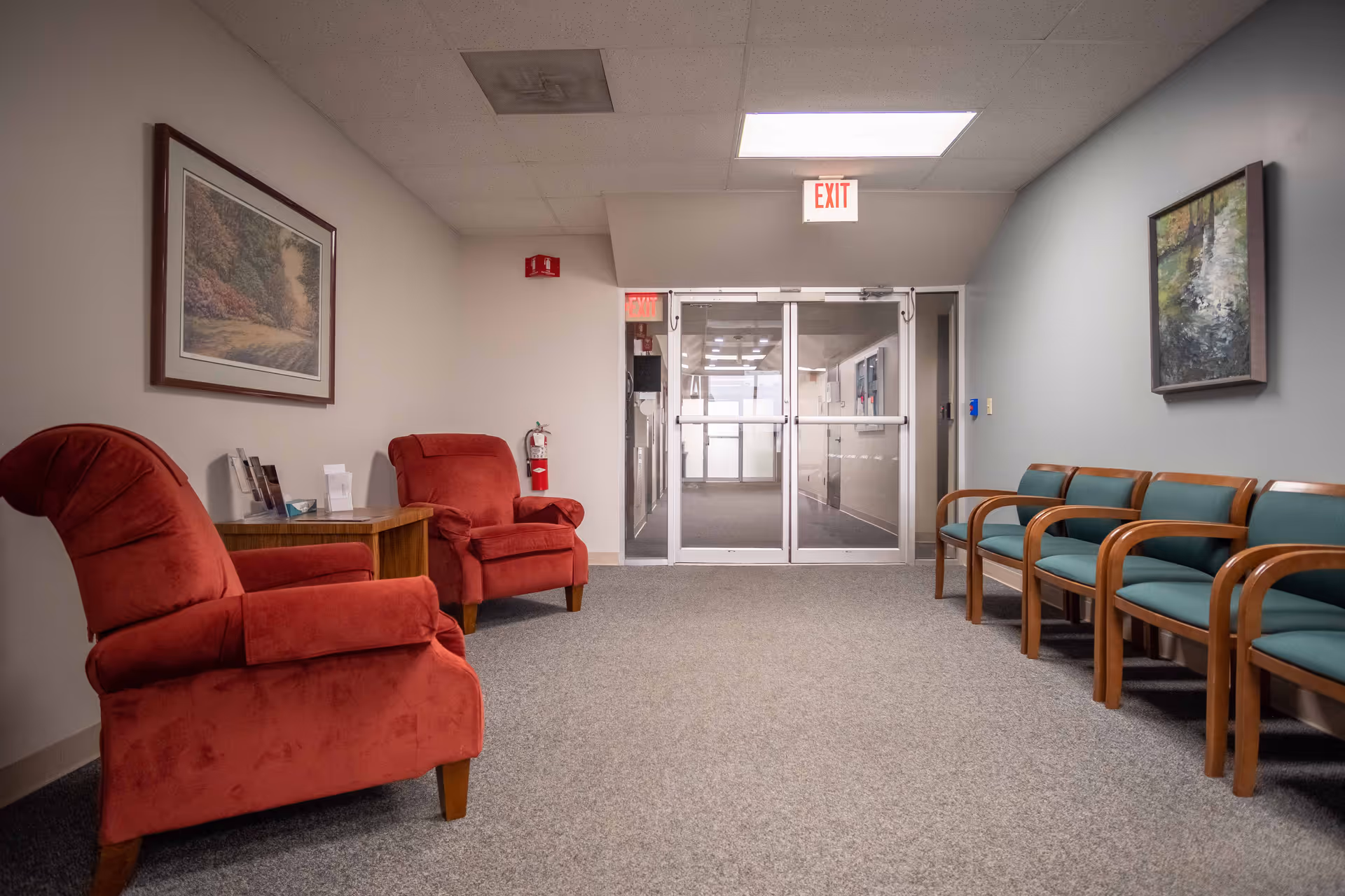 Interior waiting area with red armchairs, teal chairs, and glass doors at the back.