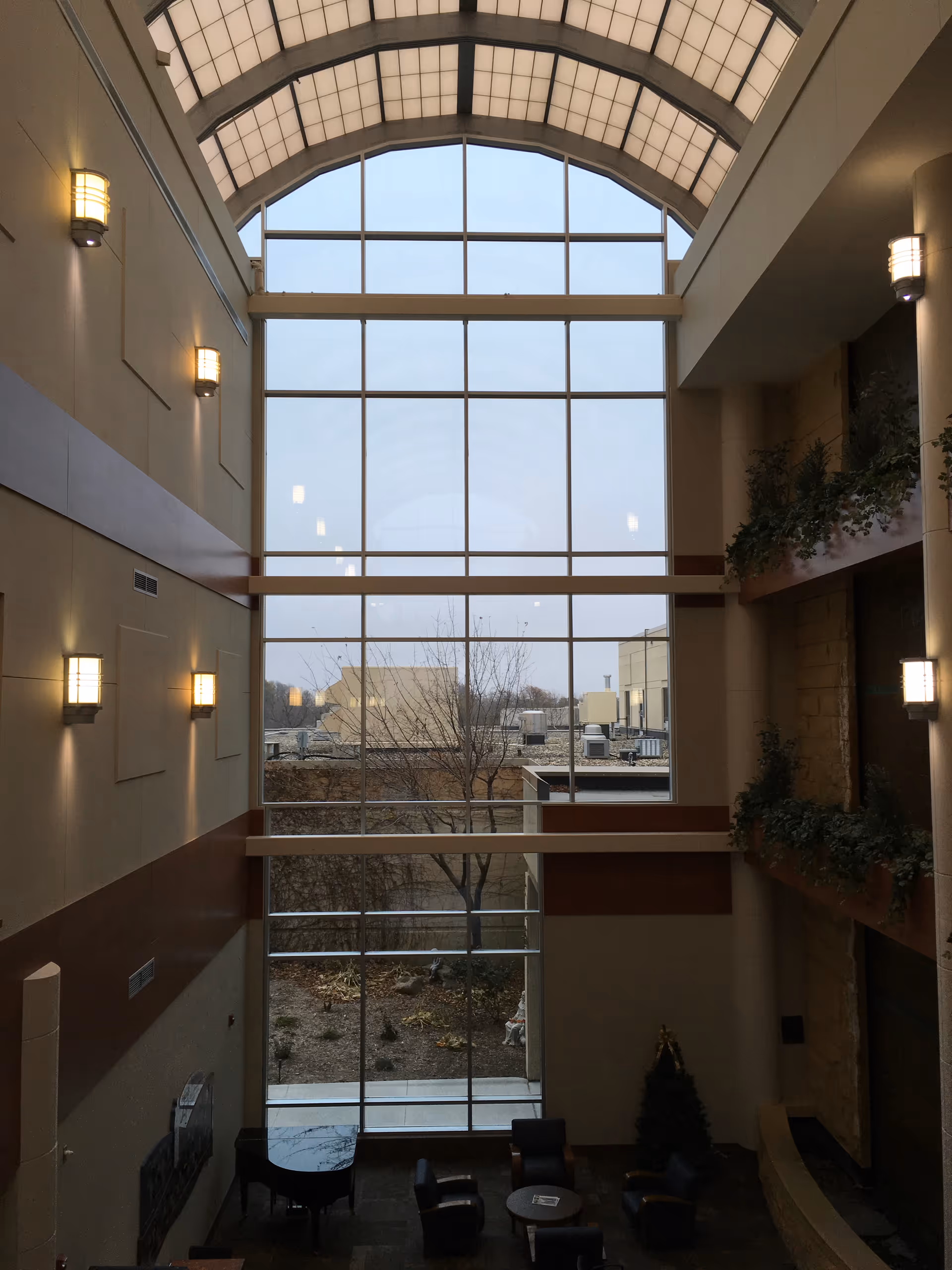 Tall glass atrium with an arched skylight, seating area, and potted plants inside.