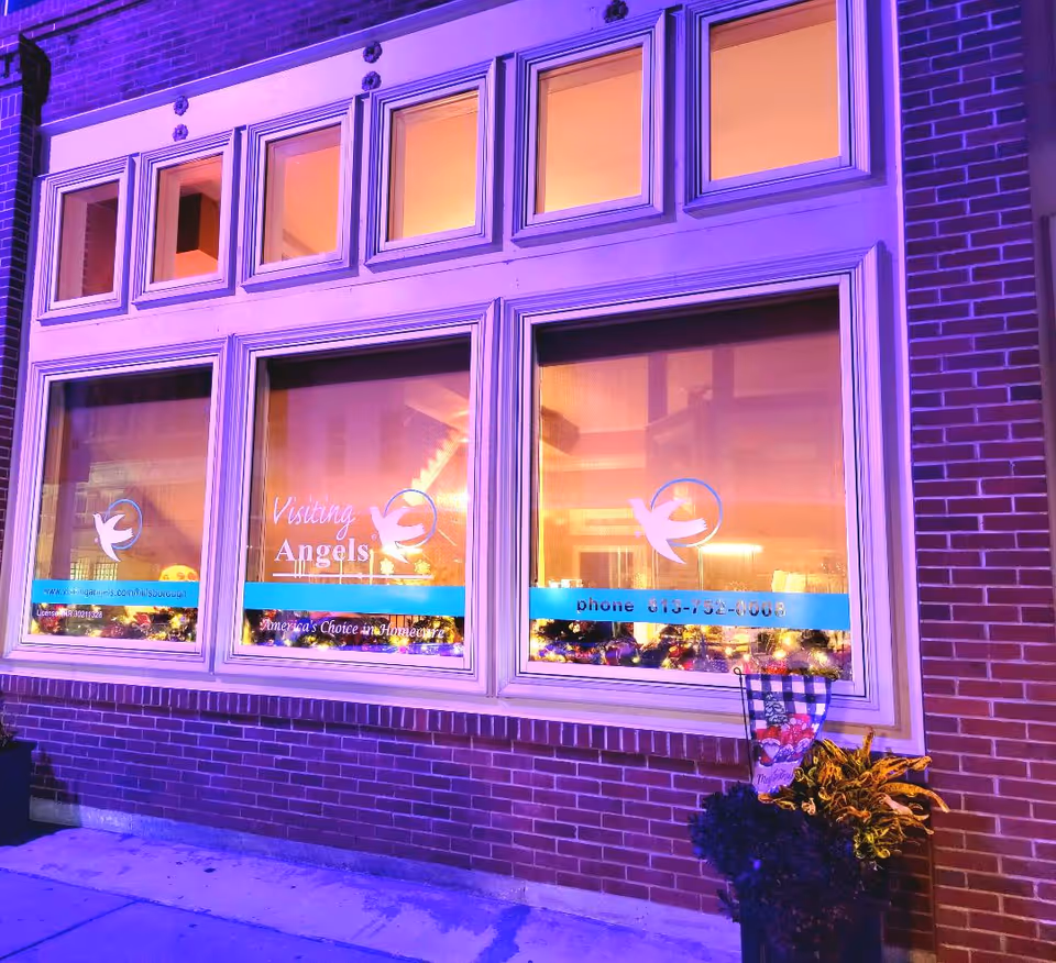 Exterior brick building with large lit windows displaying Visiting Angels logo.