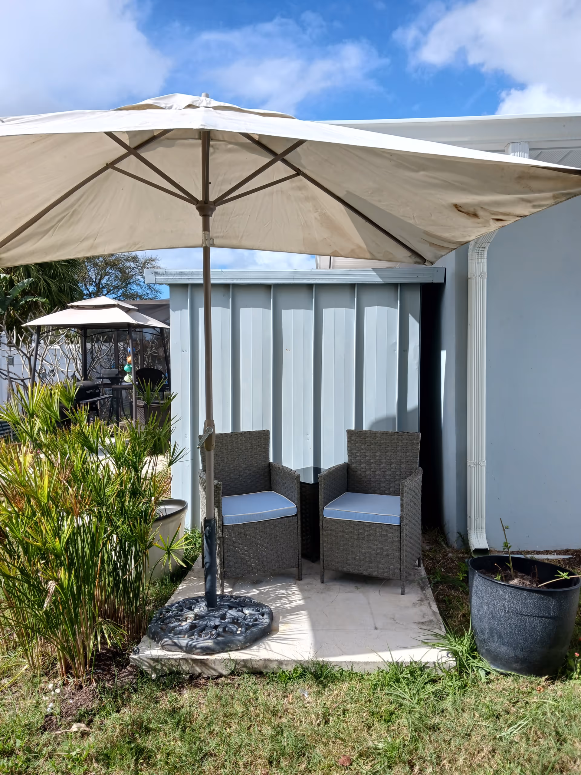 Two wicker chairs with cushions sit under a large outdoor umbrella beside a grey building.