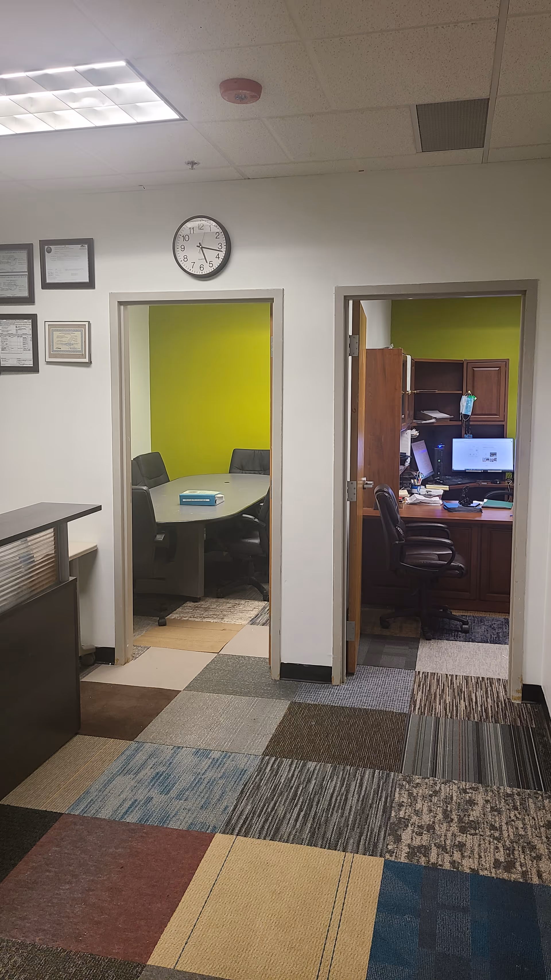Interior hallway with two doorways: green conference room, office desk area, wall clock.