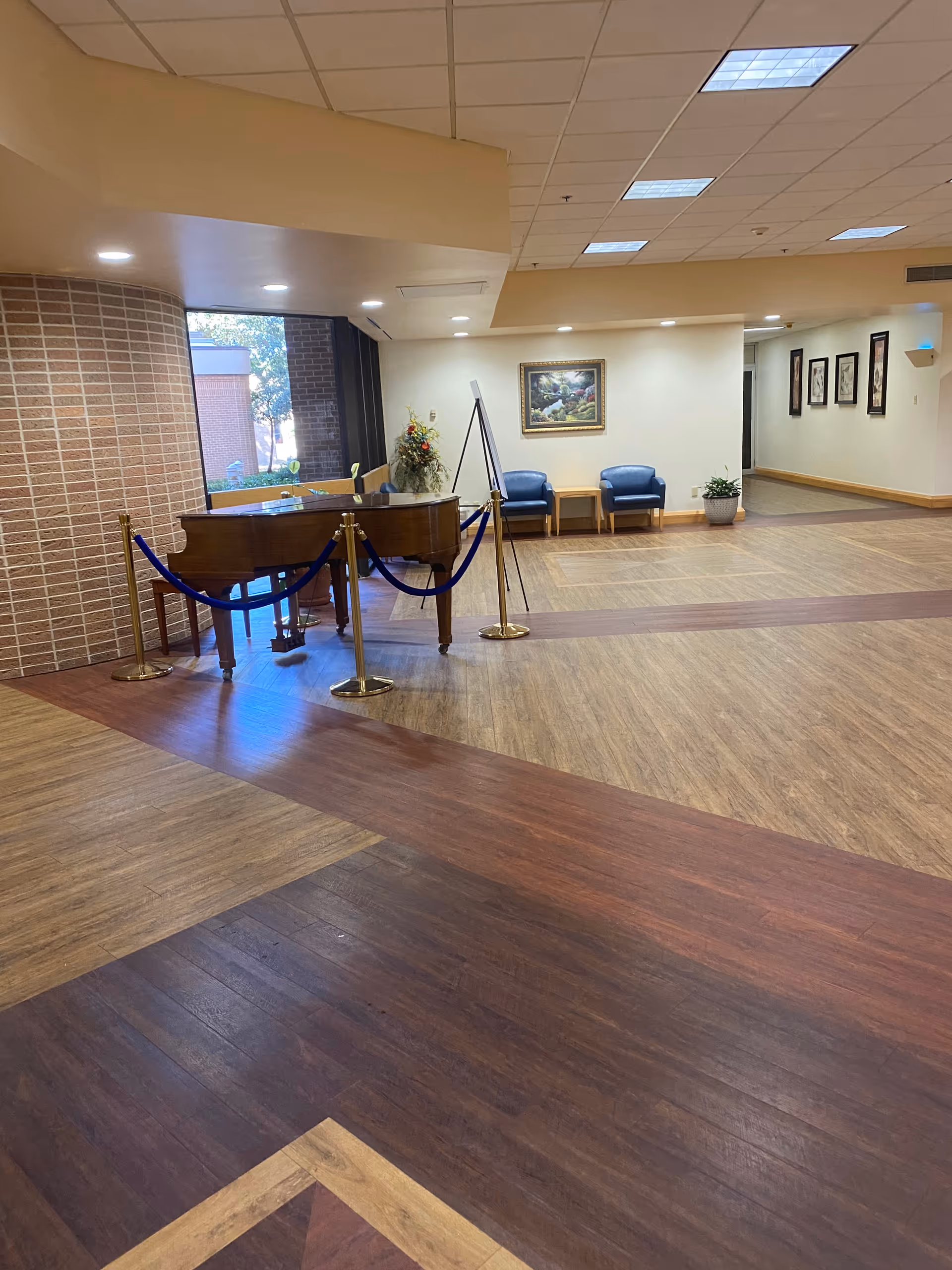 Spacious lobby area with a piano, velvet rope barriers, seating, artwork, and potted plants.