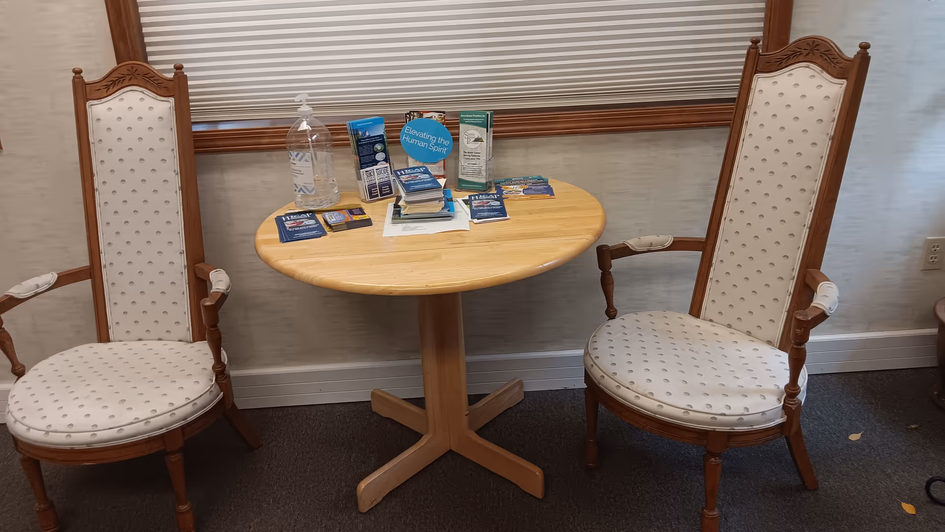 Two tall upholstered chairs face a round wooden table with brochures and hand sanitizer.